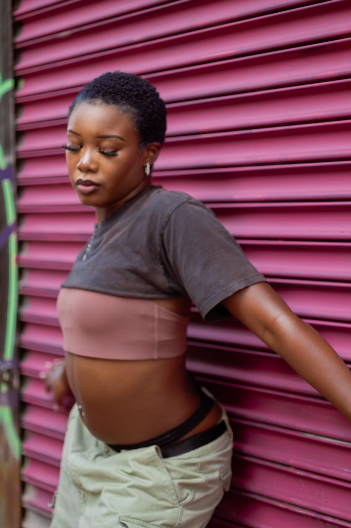 Young woman with short curly hair leaning against a pink corrugated wall, wearing a cropped top and beige pants.