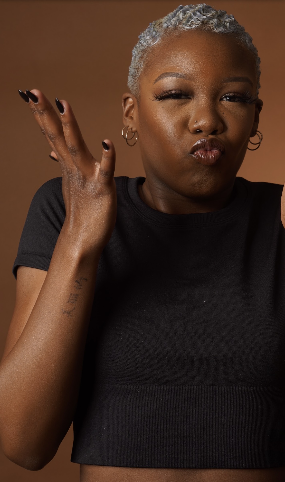 Woman with short gray hair making a playful face, lips puckered, wearing a black top, against a brown background.