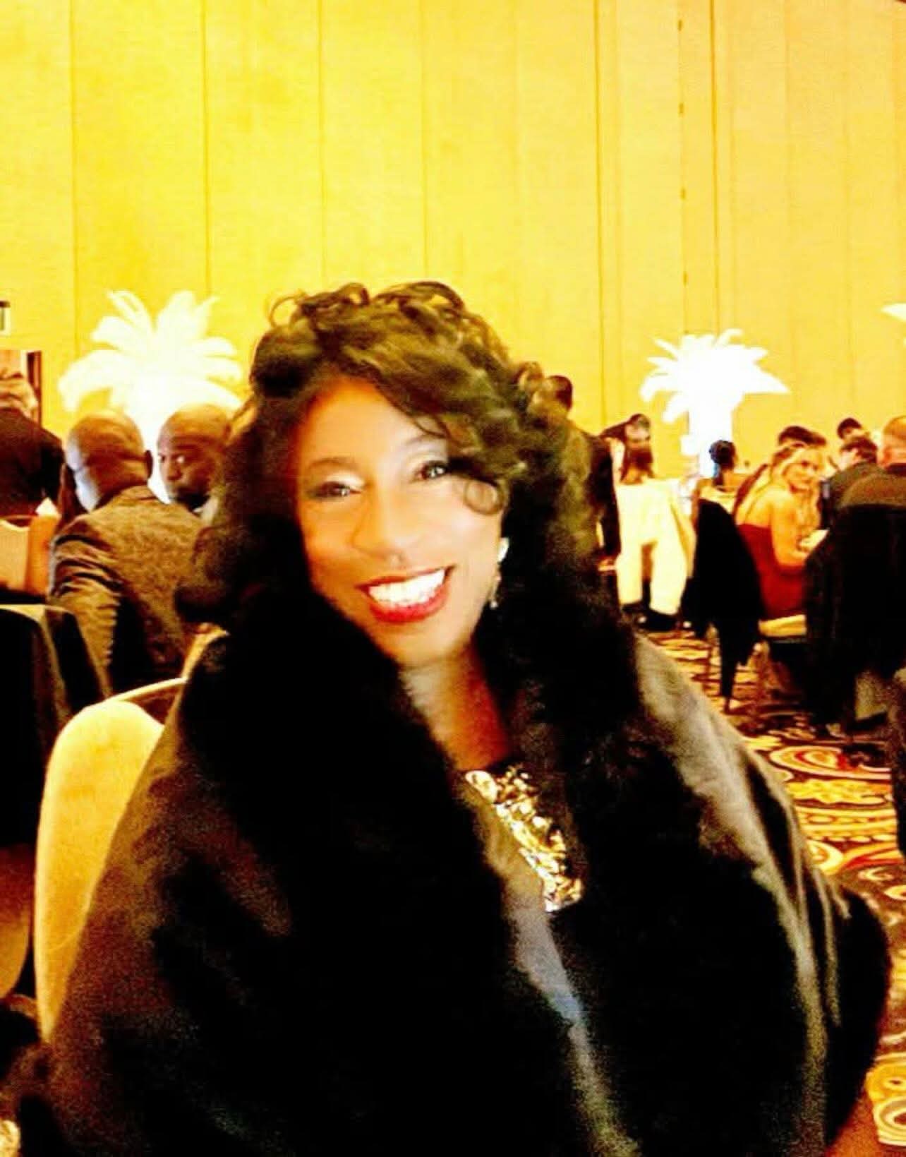 Woman with curly hair smiling, wearing a dark fur coat, at a formal event with seated guests and yellow background.
