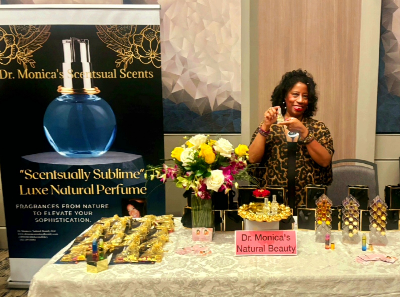 Woman with curly hair taking a selfie at a display table with perfumes and flowers, behind a sign reading 'Dr. Monica's Natural Beauty'.