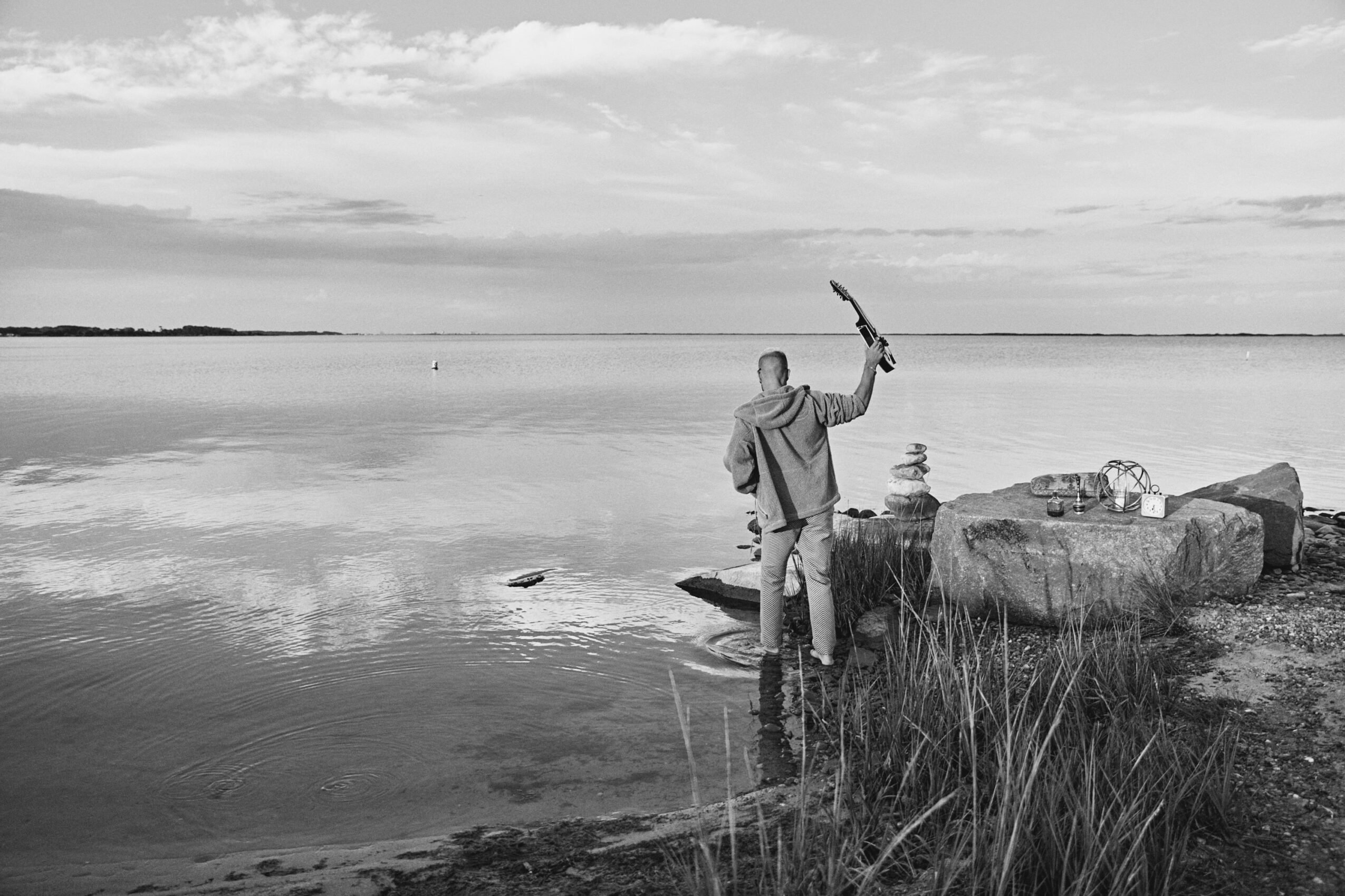Person standing on rocks by water holding a stick, with a cloudy sky and calm water in the background.