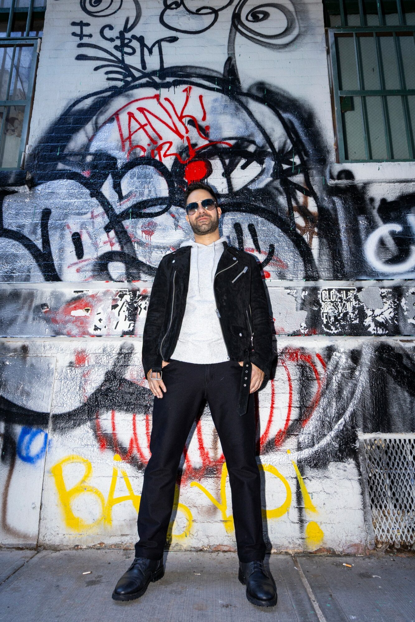 Man with sunglasses and beard in black jacket and pants standing in front of graffiti-covered wall.
