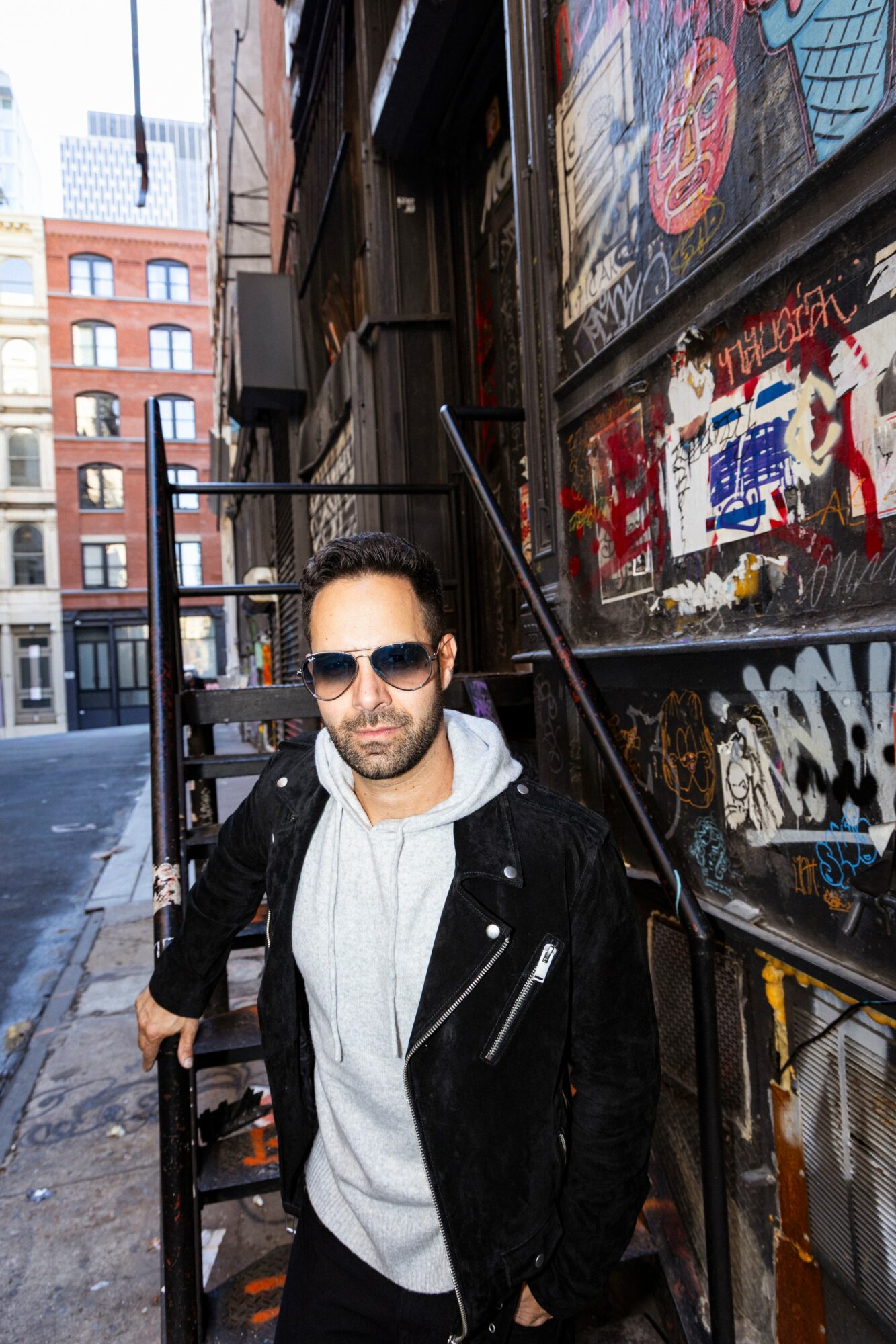 Man wearing sunglasses, hoodie, and jacket standing on city street with graffiti-covered wall behind him.