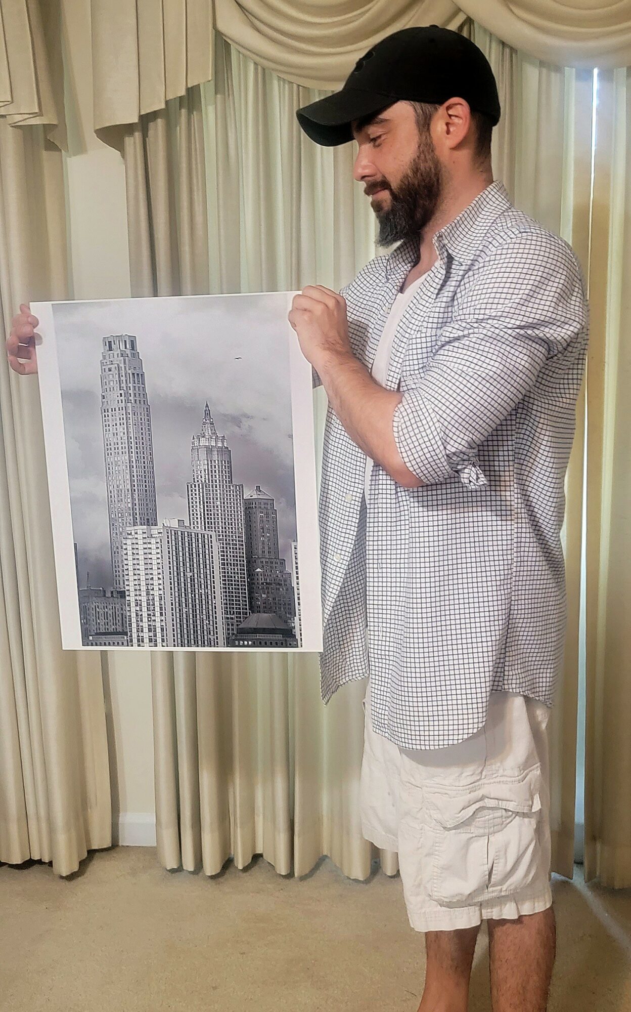 Man with beard, black cap, and casual clothing holds a black-and-white cityscape print, standing in front of curtains.
