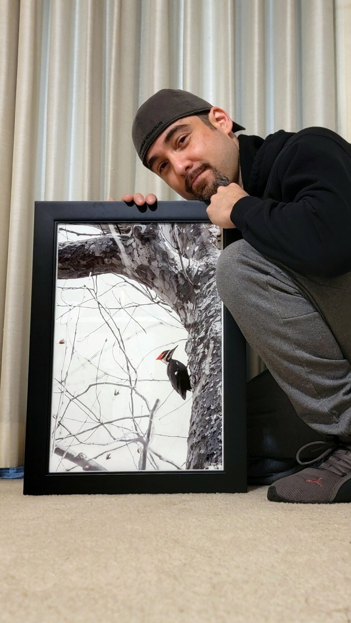 Man crouching next to a framed photograph of a snowy tree with a bird, indoors with curtains in background.