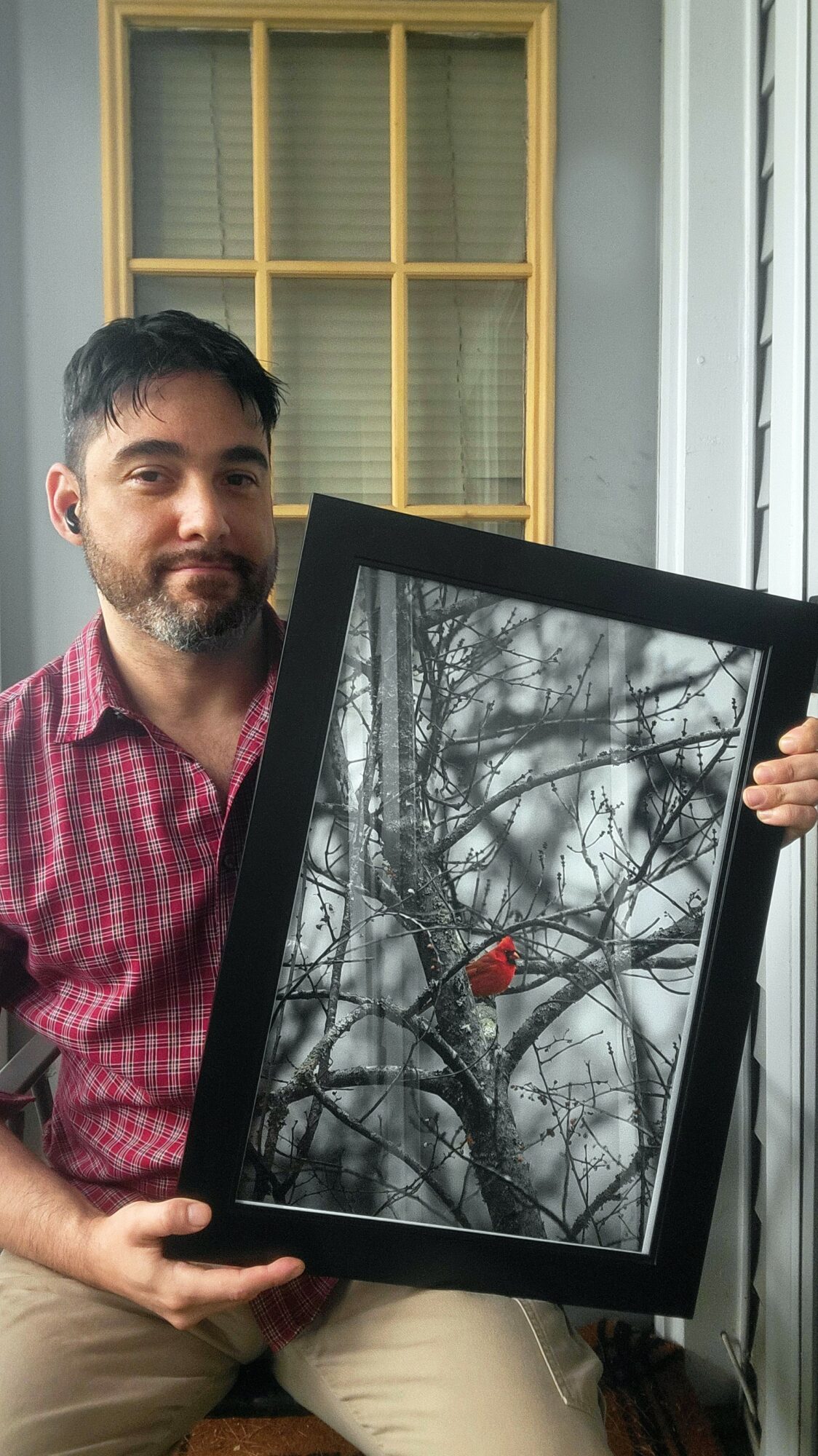Man holding a framed black-and-white photo of a bird on a tree branch, indoors near a window with yellow grid, smiling.