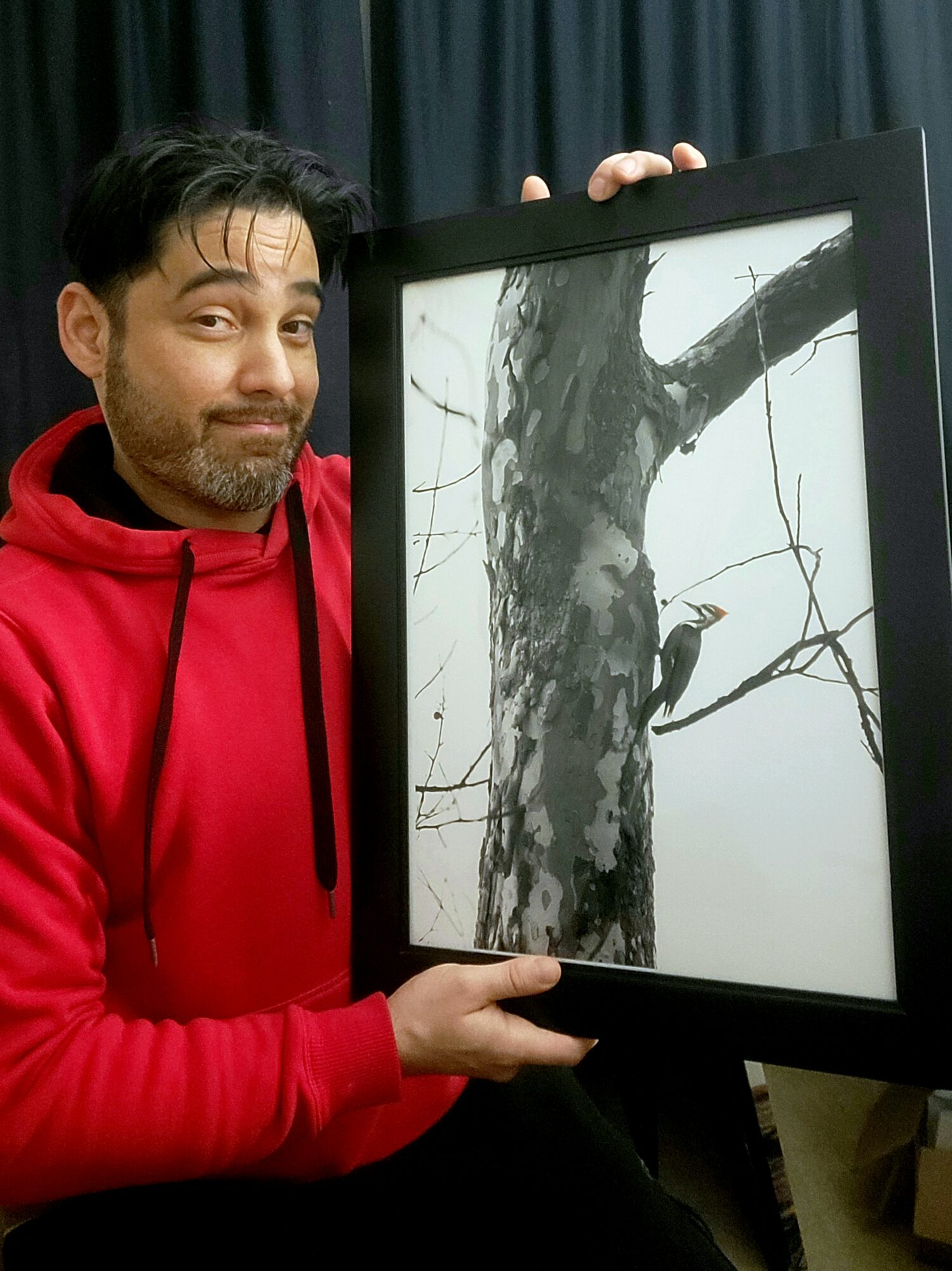 Person holding a framed black-and-white photo of a tree with a bird on a branch, smiling at the camera.