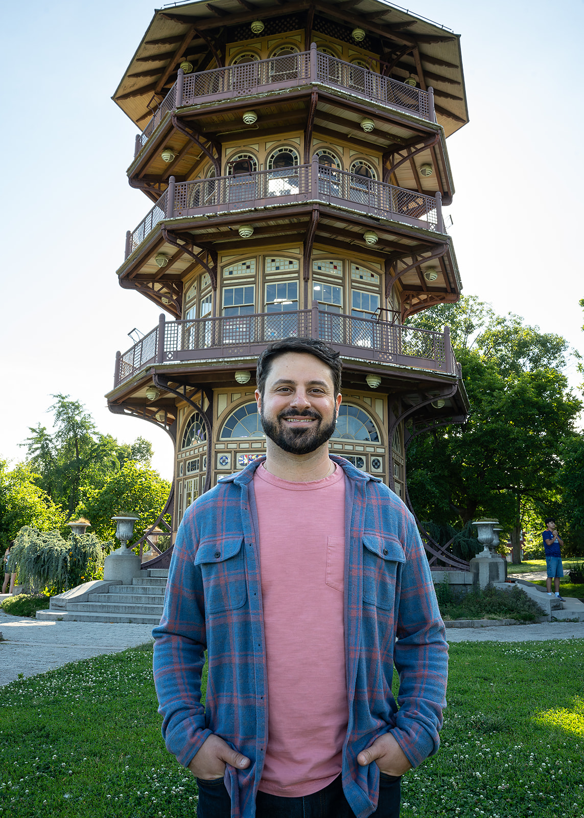 A smiling man with a beard wearing a pink shirt and blue plaid jacket stands in front of a multi-story wooden pagoda-style building with balconies, surrounded by greenery.