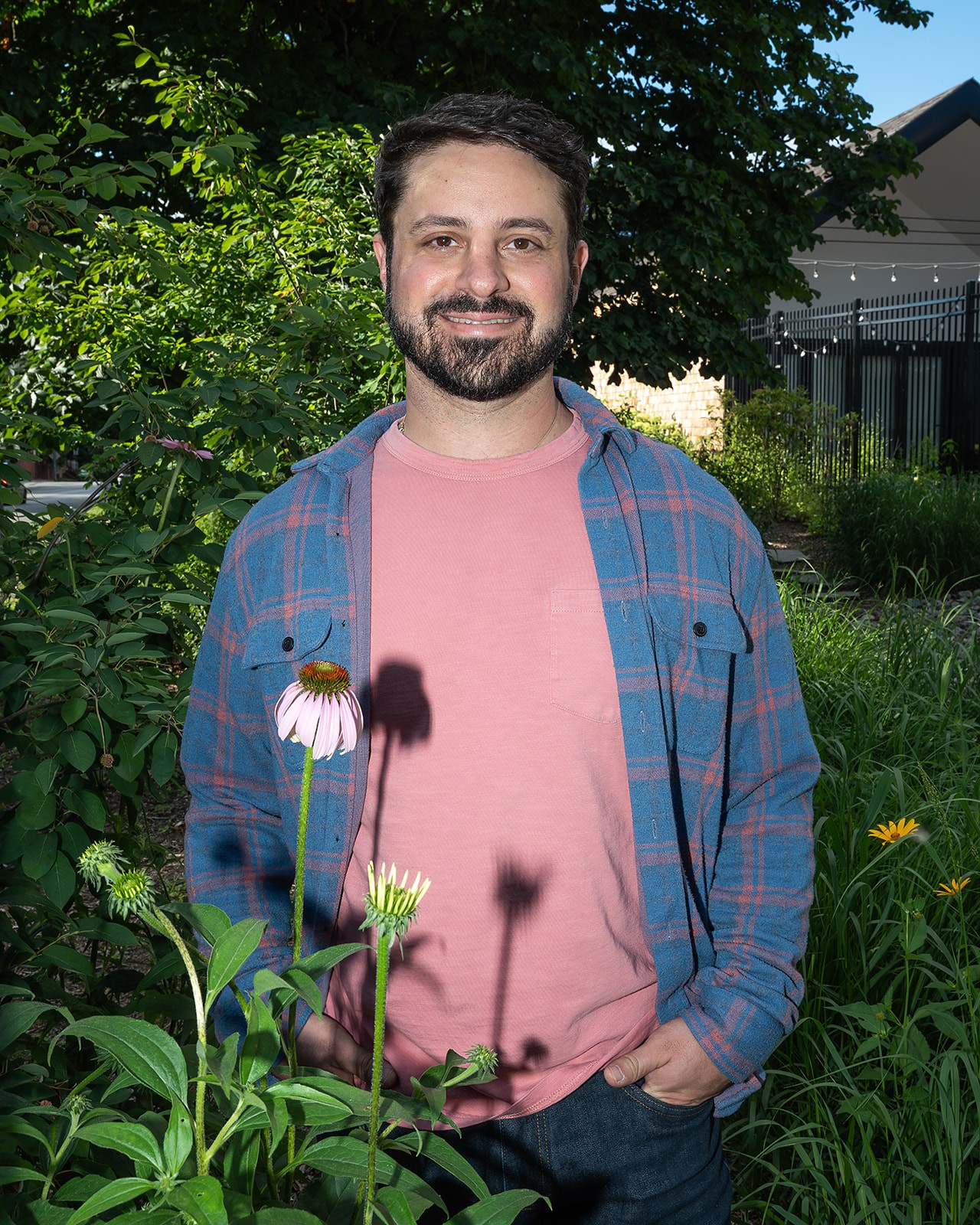 Man with beard and short dark hair smiling outdoors, wearing a pink shirt and blue plaid shirt, surrounded by greenery.