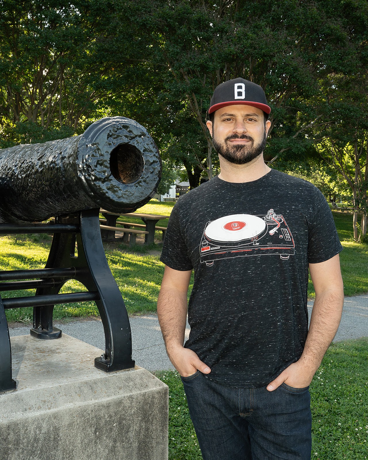Man with beard and black cap standing outdoors next to a cannon, smiling, with trees and a park in the background.