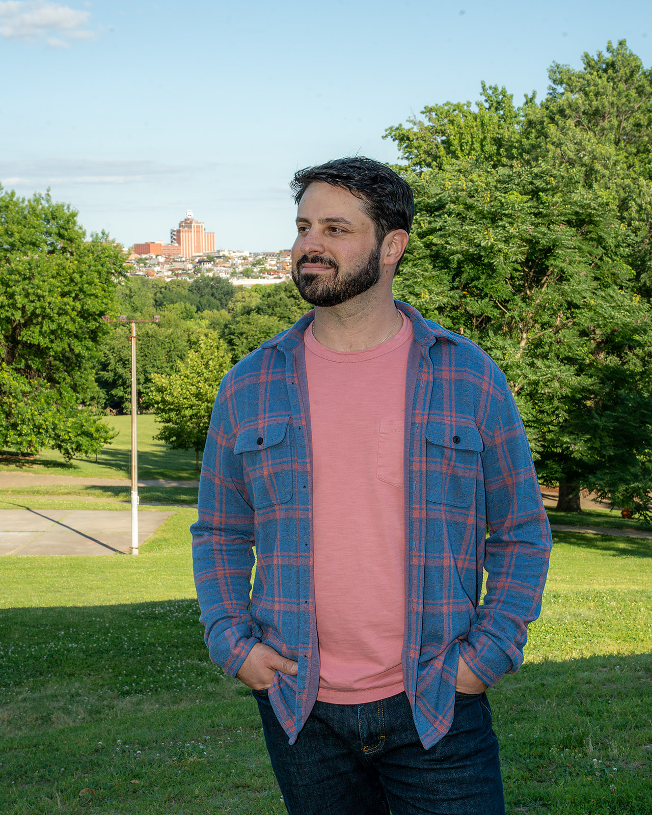 Man with dark hair and beard smiling outdoors, wearing a pink shirt and blue plaid shirt, in a park with trees and city in background.