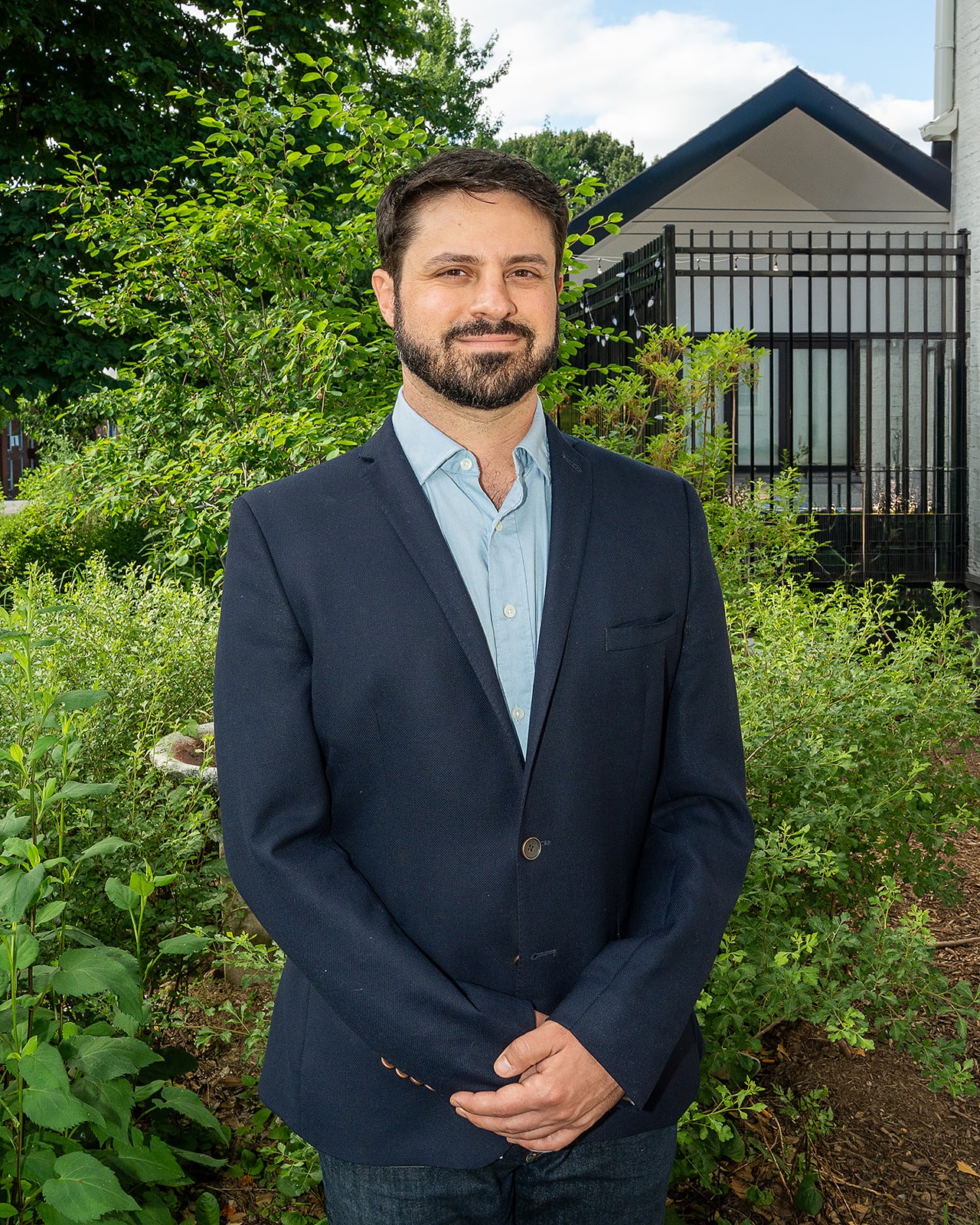 Man in a navy suit and light blue shirt standing outdoors with greenery and a house in the background.