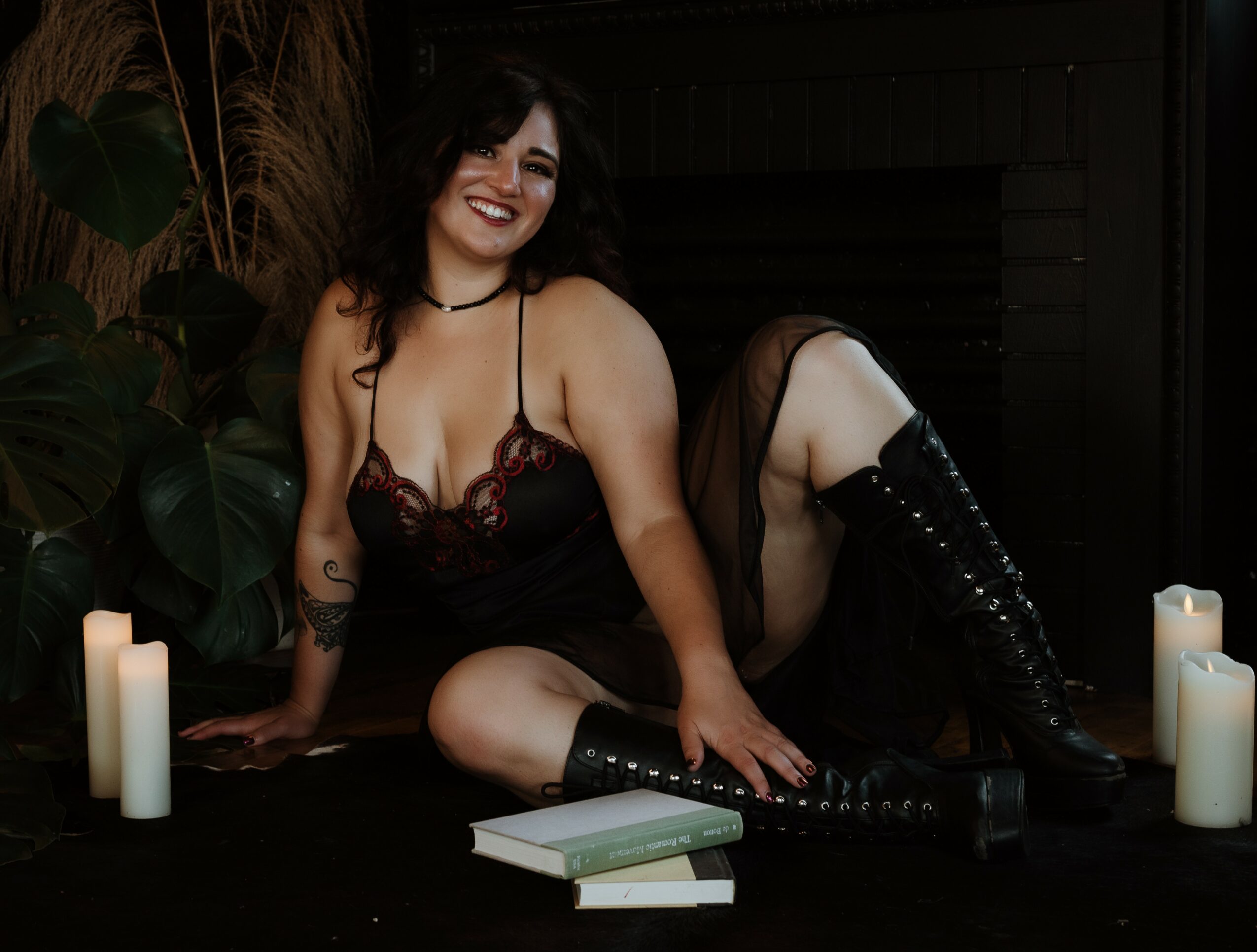 Woman with dark hair smiling, sitting on the floor surrounded by candles and books, wearing a black dress and boots.
