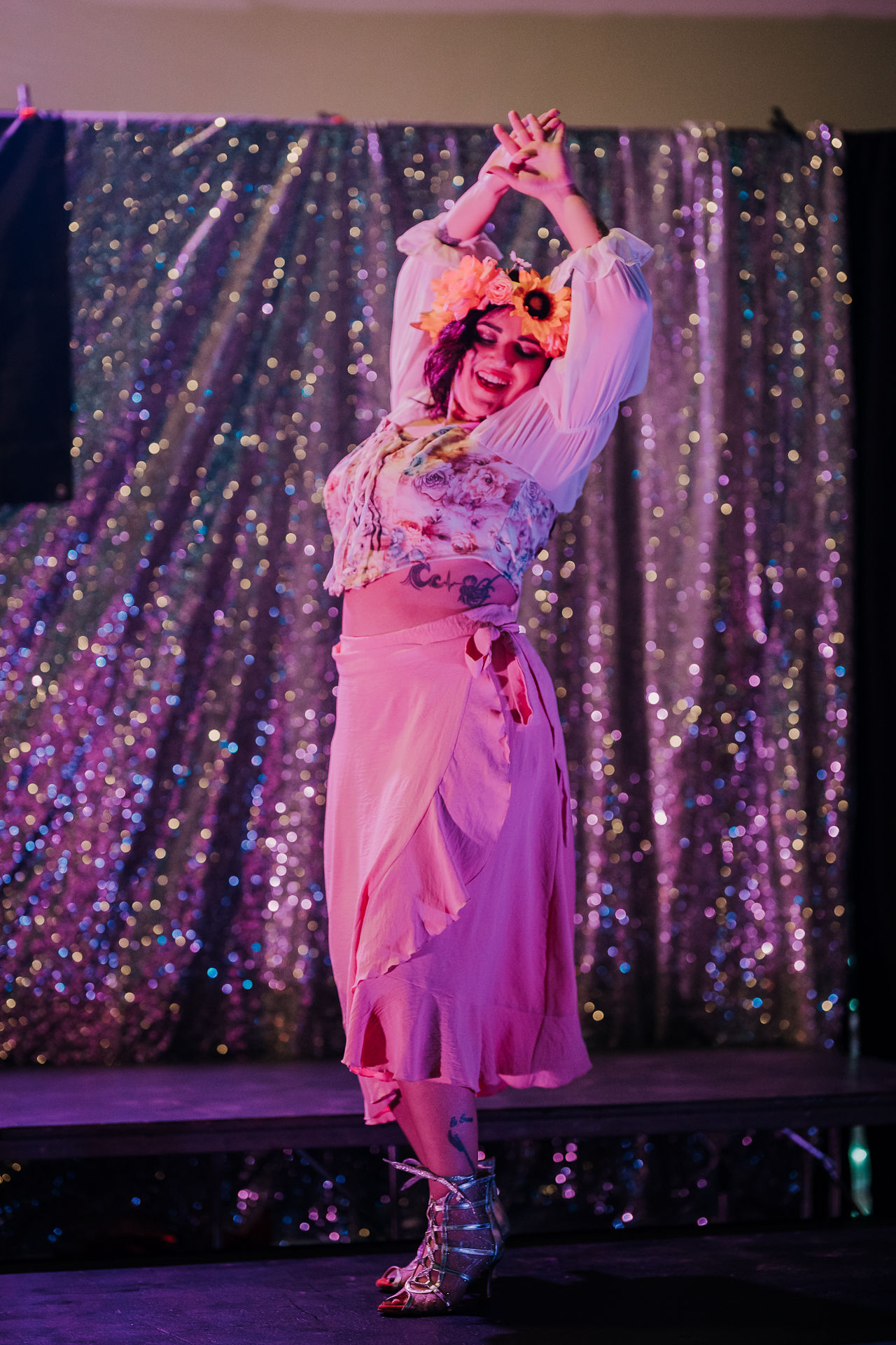 Person in colorful costume with a wig, dancing on stage with a sparkly curtain background.