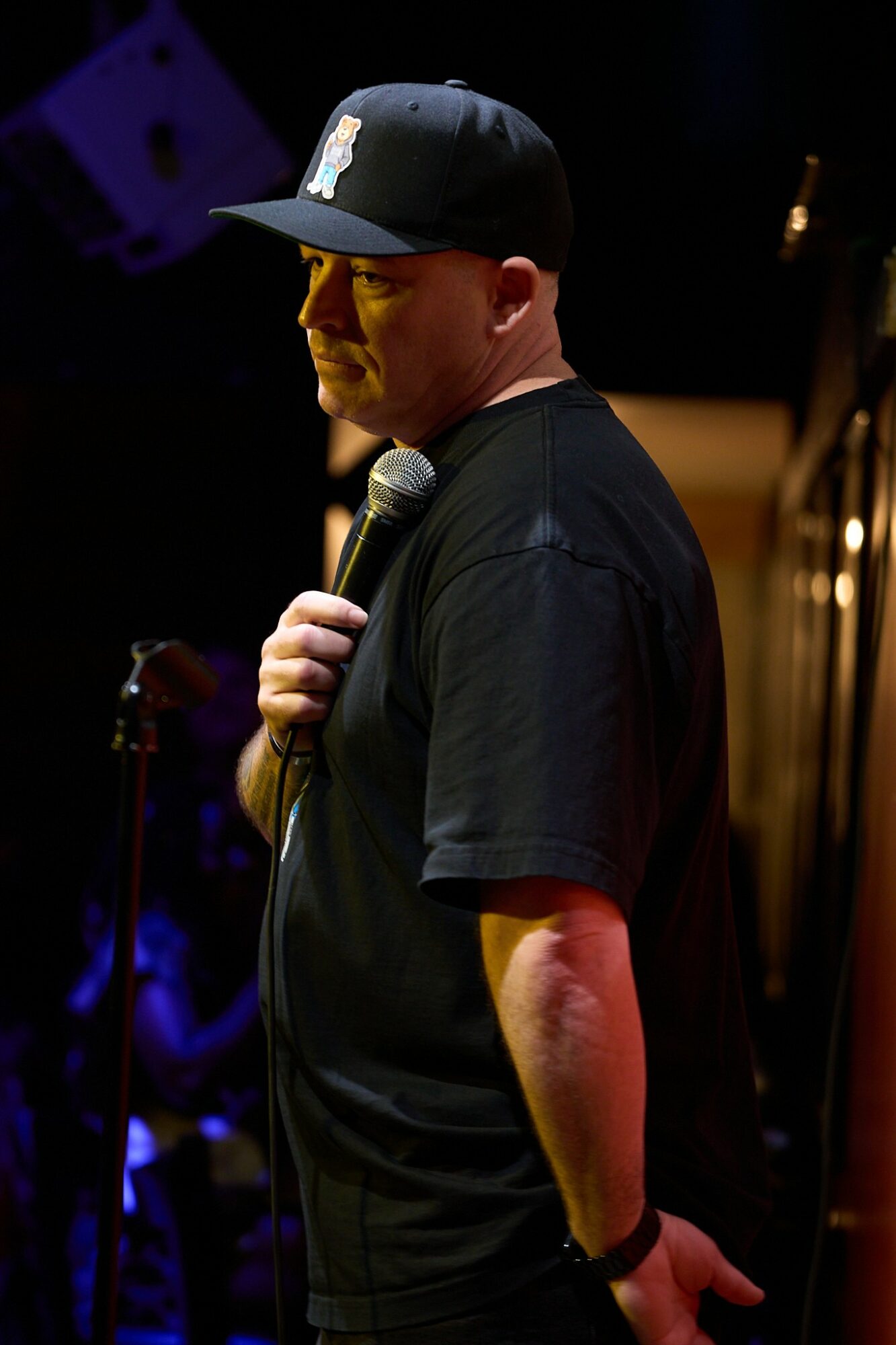 Person holding a microphone on stage, wearing a black cap and T-shirt, with a dark background.