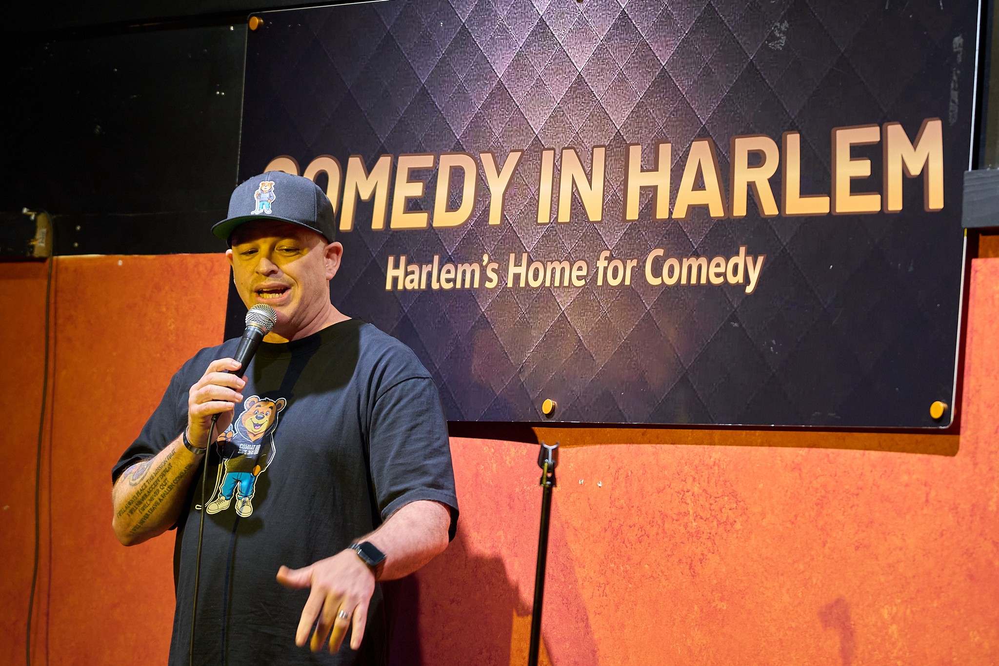 Person wearing a hat and glasses holding a microphone on stage with a sign that reads 'COMEDY IN HARLEM' behind them.