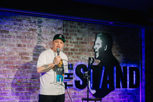Person holding microphone on stage with brick wall background and text reading 'THE STAND'.