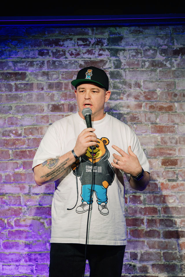 Person standing on stage with brick wall background, holding microphone, wearing a cap and graphic t-shirt, speaking.
