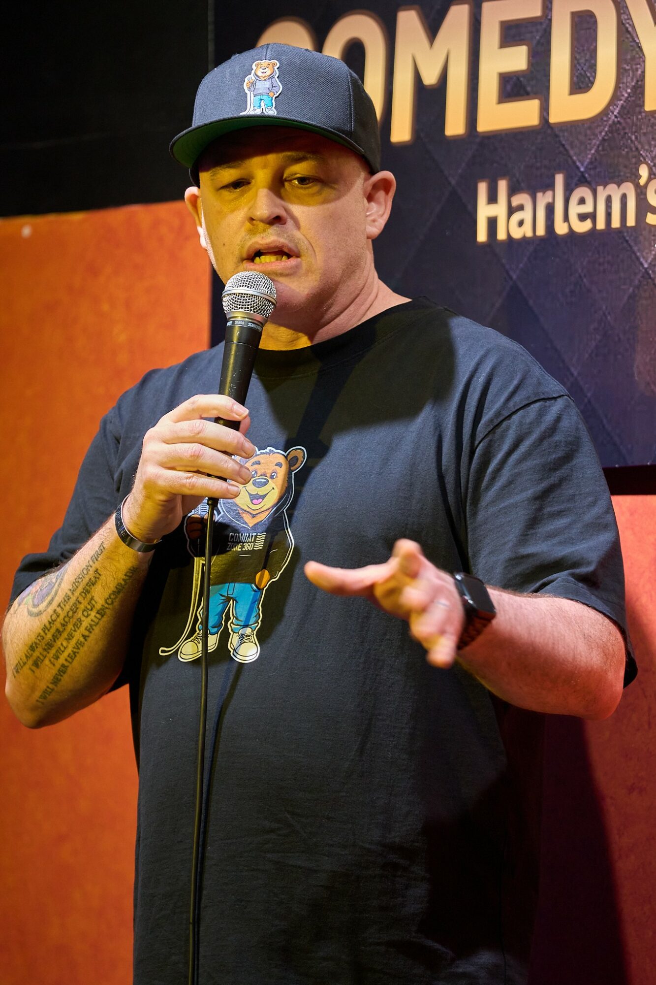 Man wearing a baseball cap and black T-shirt holding a microphone, speaking on stage with a sign behind him.