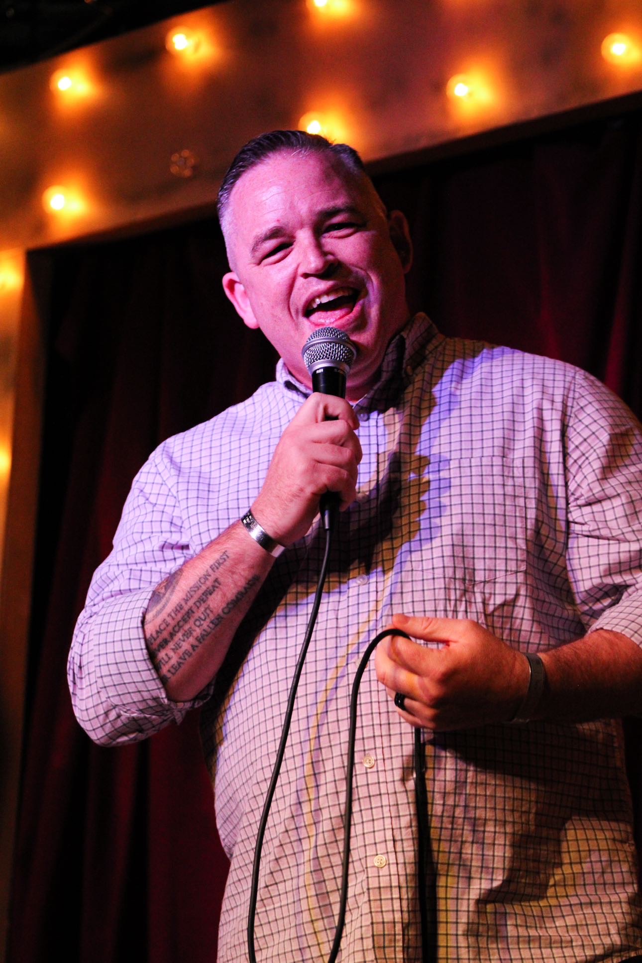 Person holding microphone, smiling, performing on stage with warm lighting and dark curtain background.