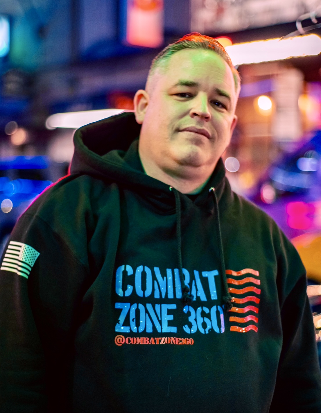 Young man in black hoodie with colorful text and graphics, illuminated city background at night.