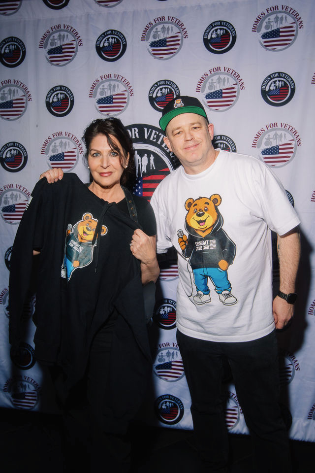 Two people standing in front of a backdrop with American flags and logos, smiling and posing for the photo.