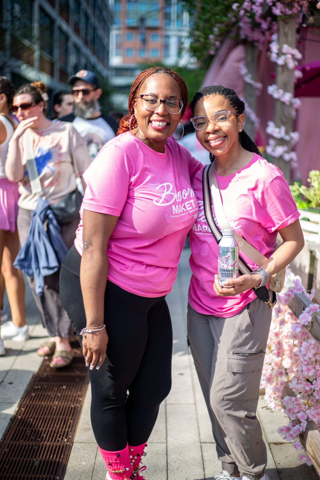 Two women smiling outdoors, wearing pink shirts, with a group of people and pink flowers in the background.