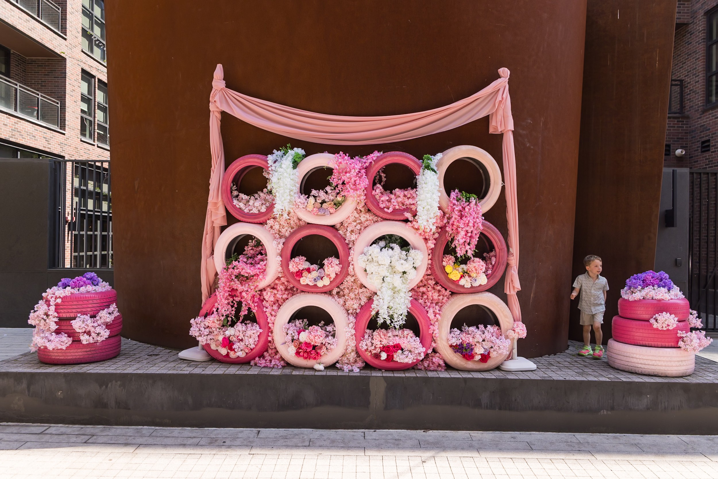 Pink and white floral display with circular frames, draped pink fabric, and two cake-like decorations on each side, outdoors.