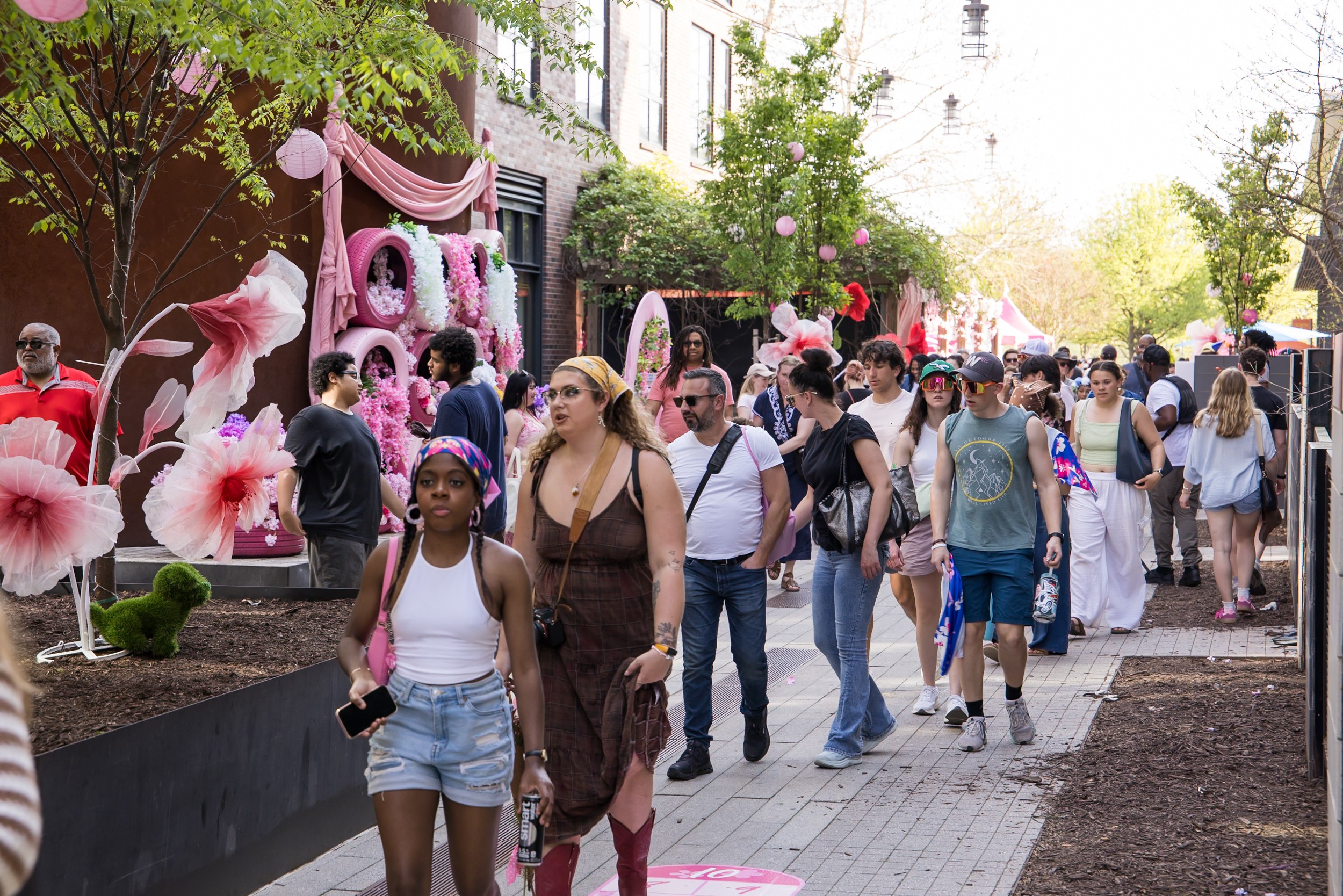 Crowd of people walking on a sidewalk during a festive outdoor event with pink decorations and trees.