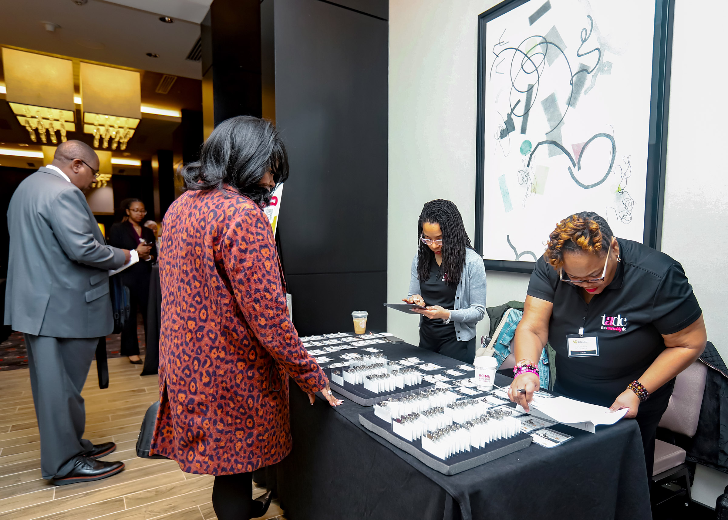 People at a registration table with two women behind it, one using a tablet, in a modern indoor setting.
