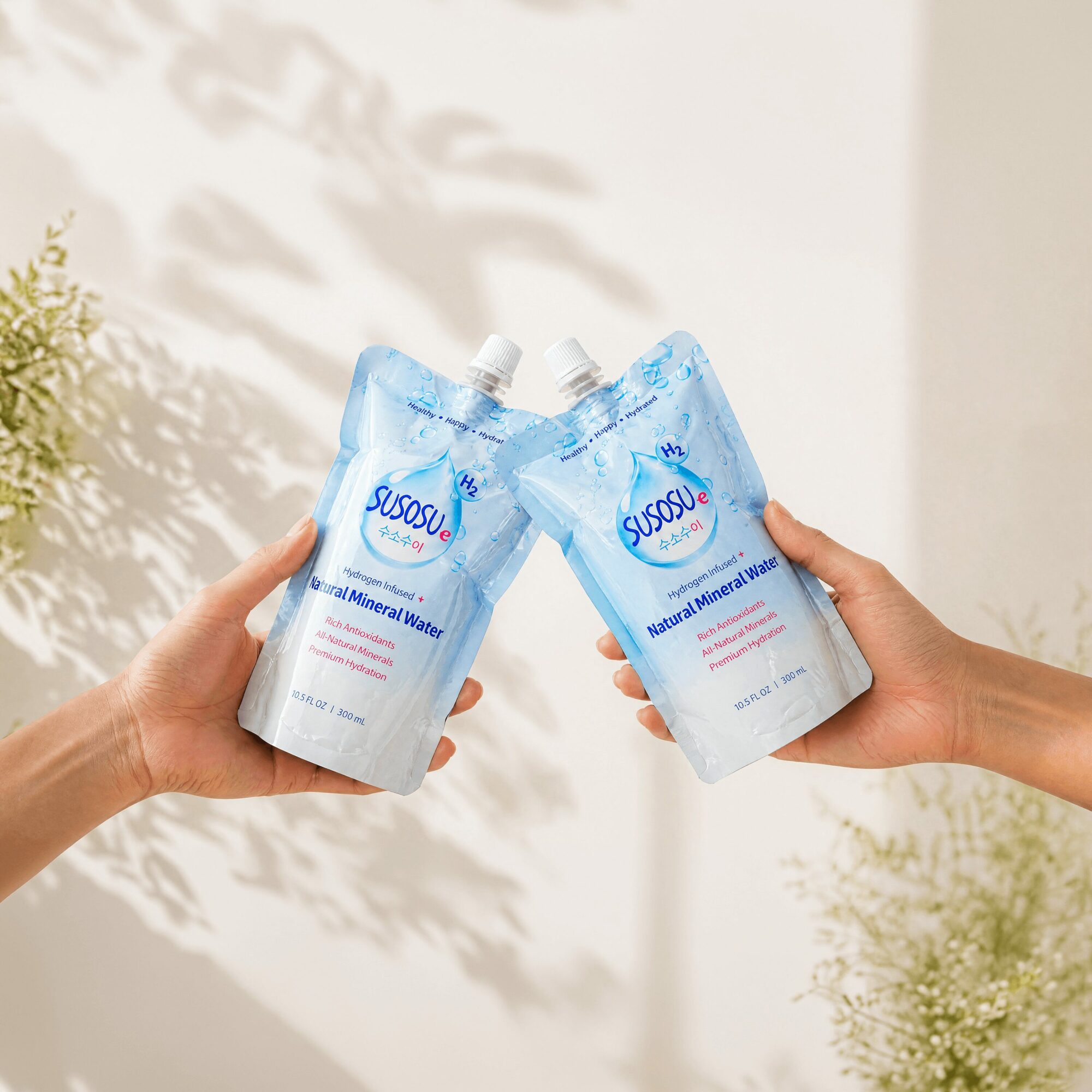 Two hands holding identical blue and white pouches with spouts, against a light background with shadows and plants.