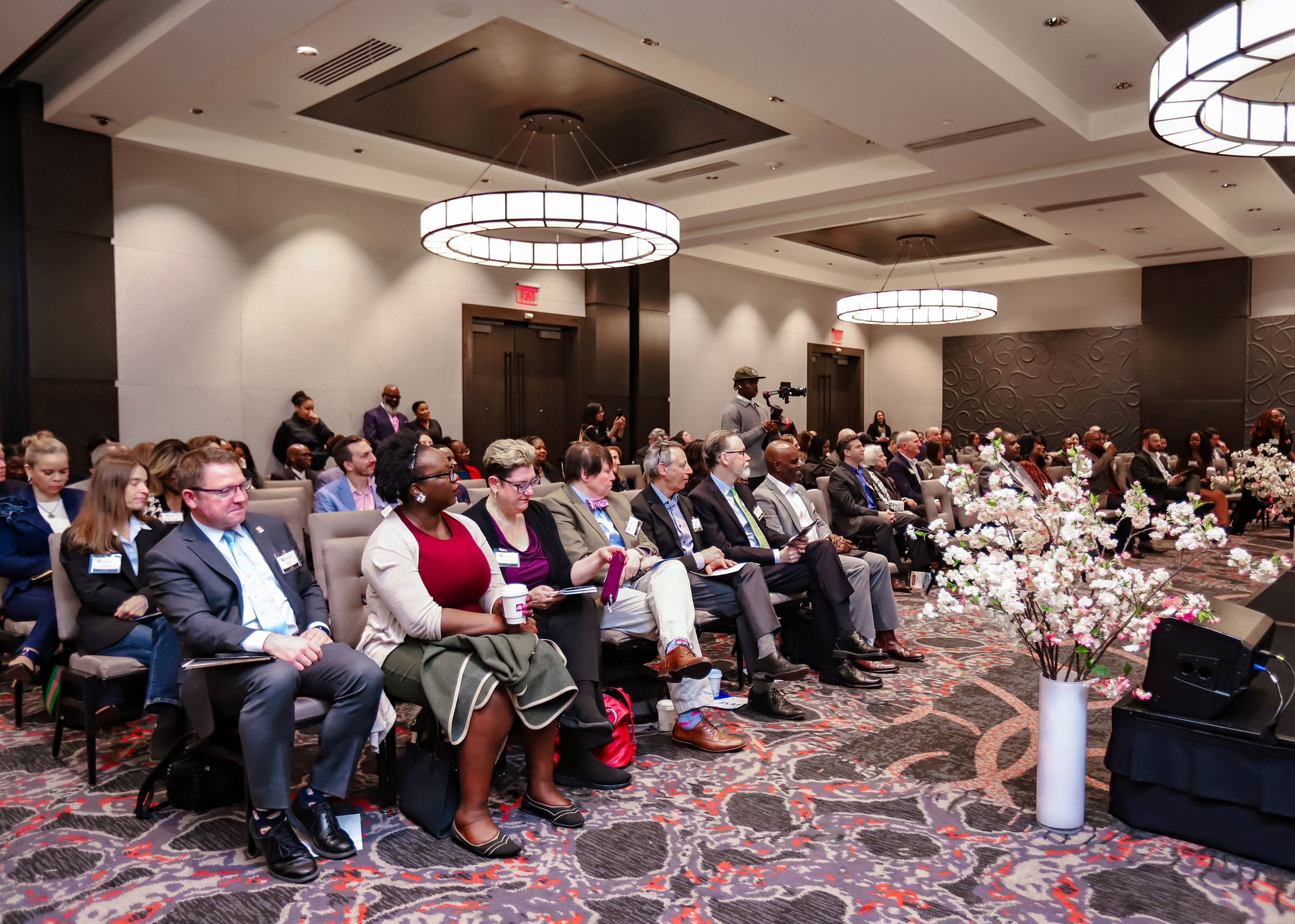 Audience seated in a conference room with floral arrangements and large circular ceiling lights.