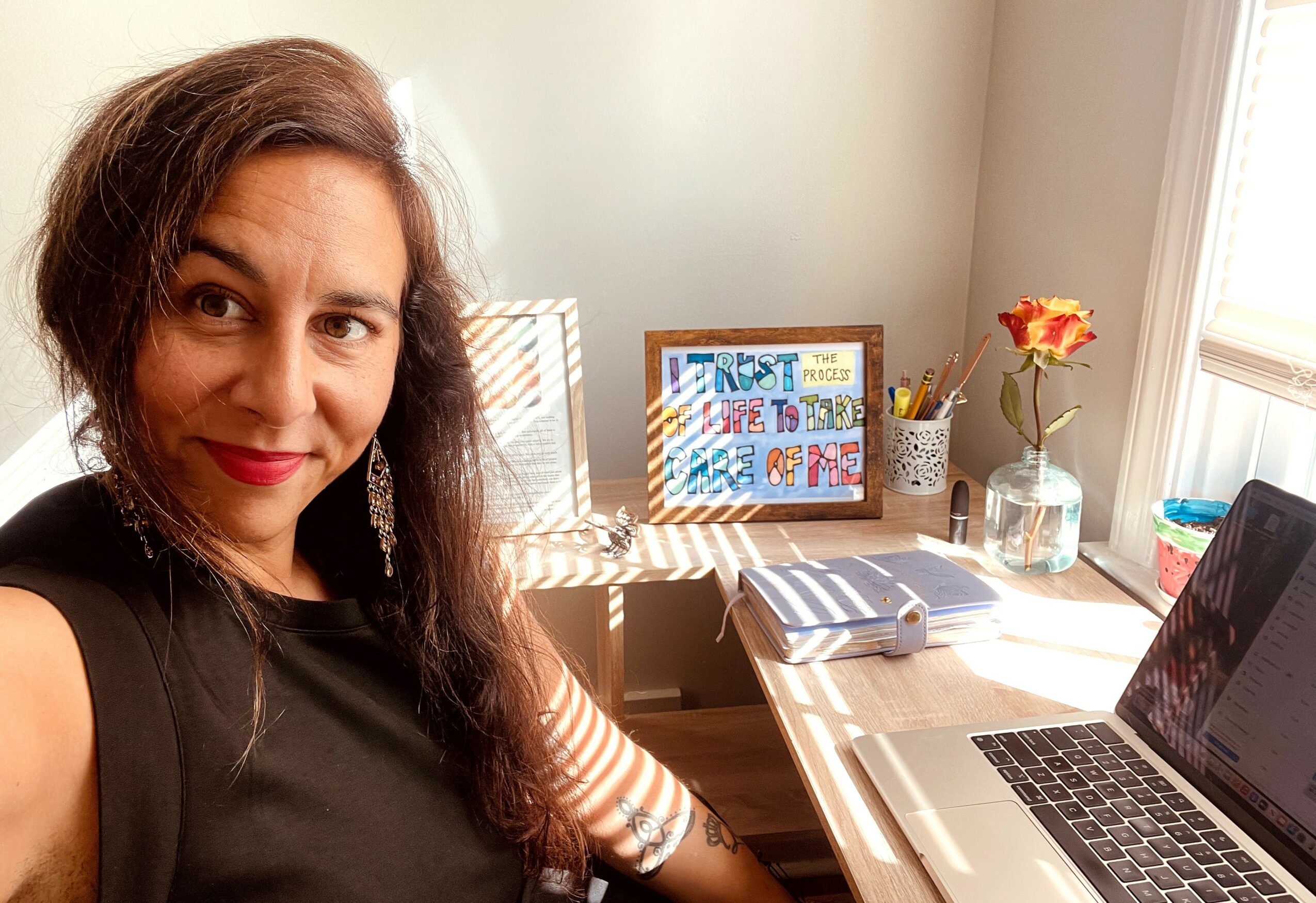 Woman with long dark hair sitting at a desk with a laptop, colorful artwork, and a vase with flowers on a windowsill.
