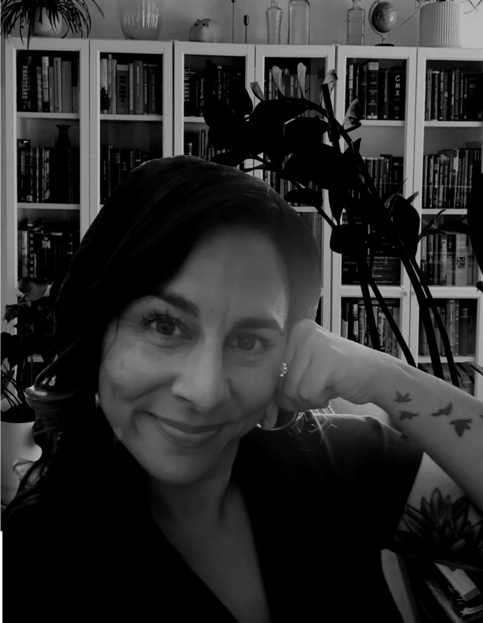 Woman with dark hair smiling, resting her head on her hand, in front of a bookshelf with plants, in black and white.