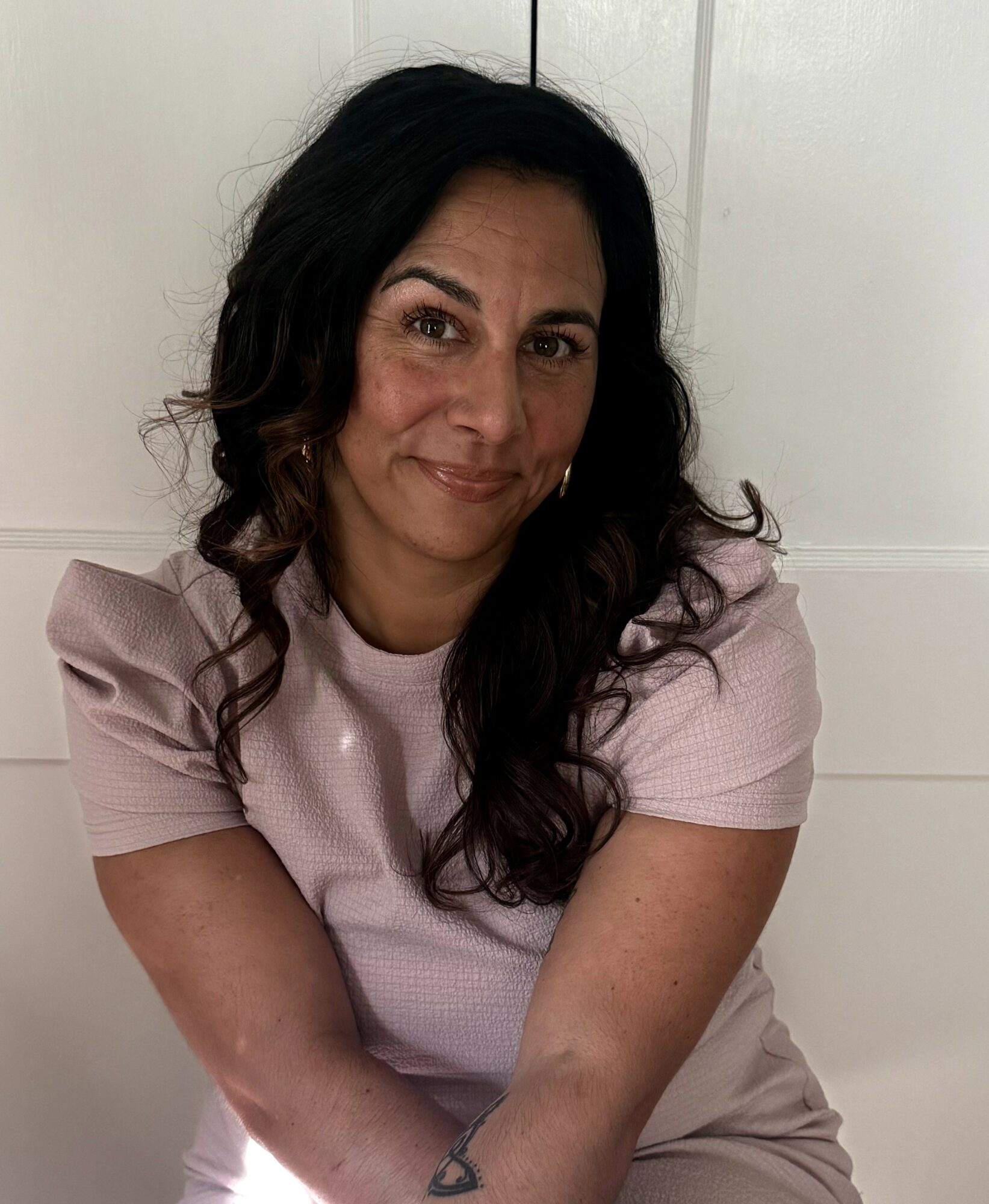 Woman with dark wavy hair smiling, wearing a light pink top, sitting against a white background.