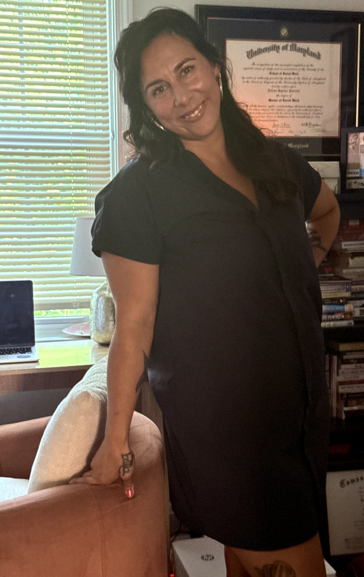 Woman with dark hair smiling, wearing a black dress, standing indoors near a window with blinds, bookshelf, and framed certificate behind her.