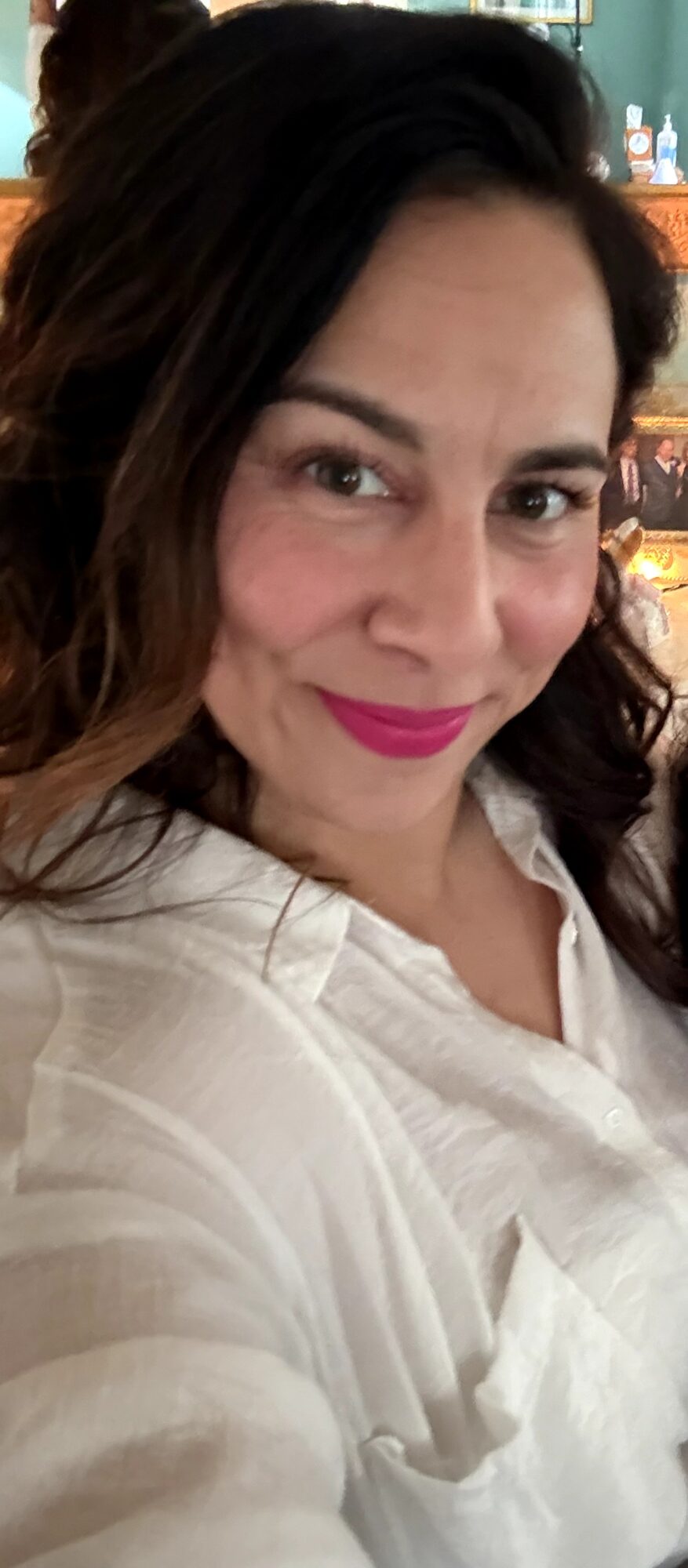 Close-up of a woman with dark hair, smiling, wearing a white shirt, in an indoor setting.
