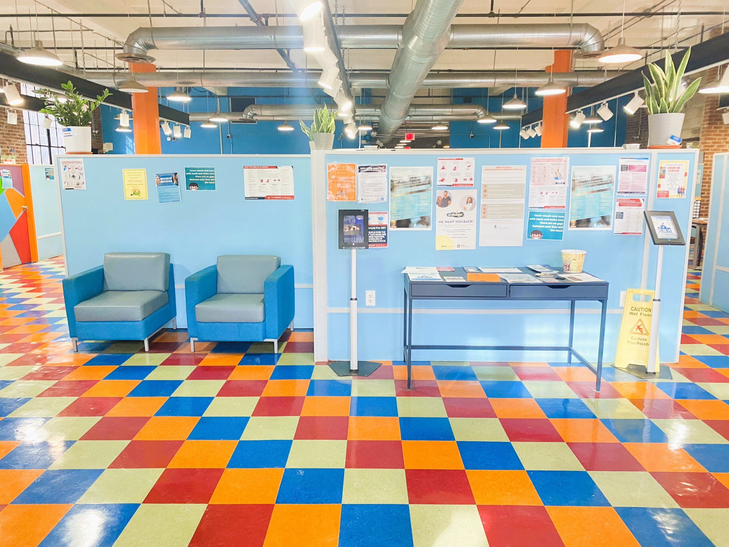 Indoor space with blue chairs, display boards, and a table, colorful tiled floor, and ceiling pipes.