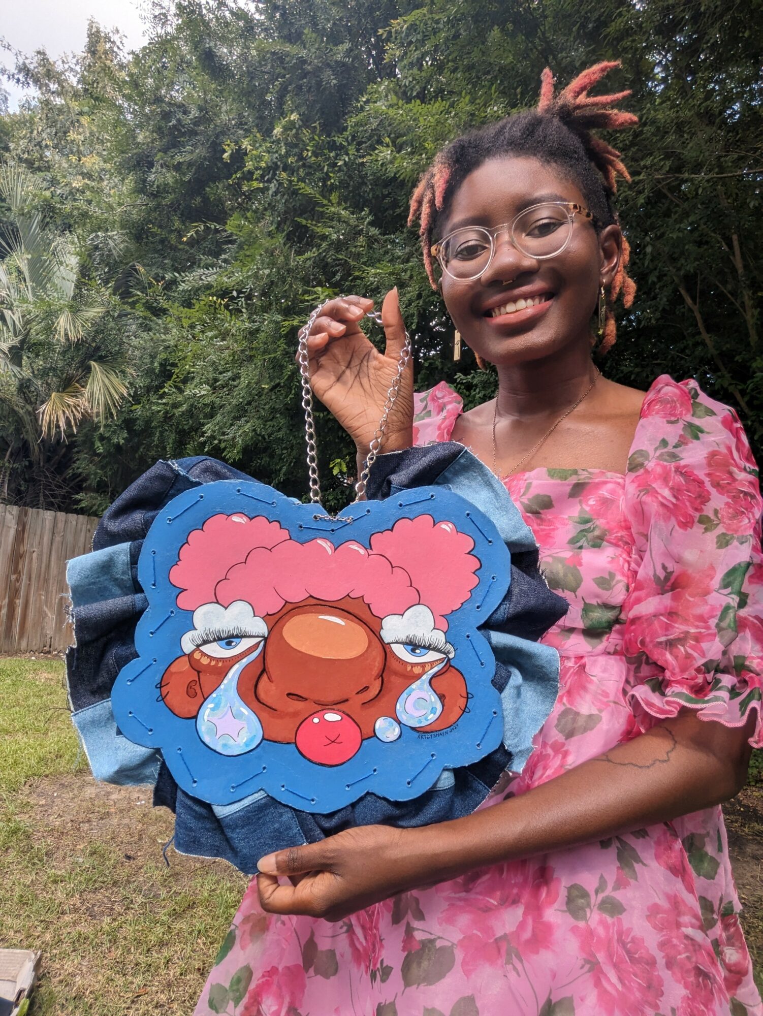 Young girl with glasses and a pink floral dress holding a colorful bag with a cartoon face, outdoors with trees in background.