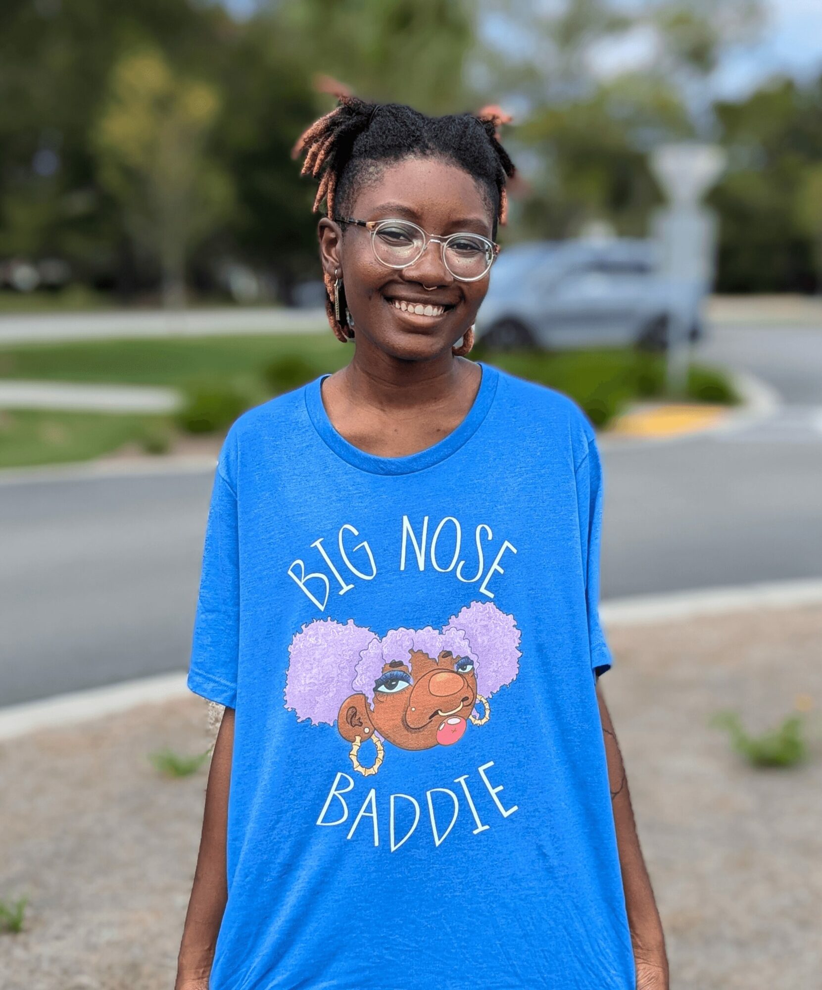 Smiling girl with glasses and pigtails outdoors, wearing a blue T-shirt with a cartoon face and text.