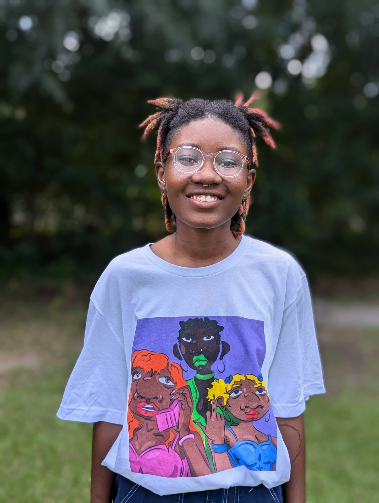 Smiling young woman with glasses and styled hair outdoors, wearing a graphic T-shirt with colorful illustrations.