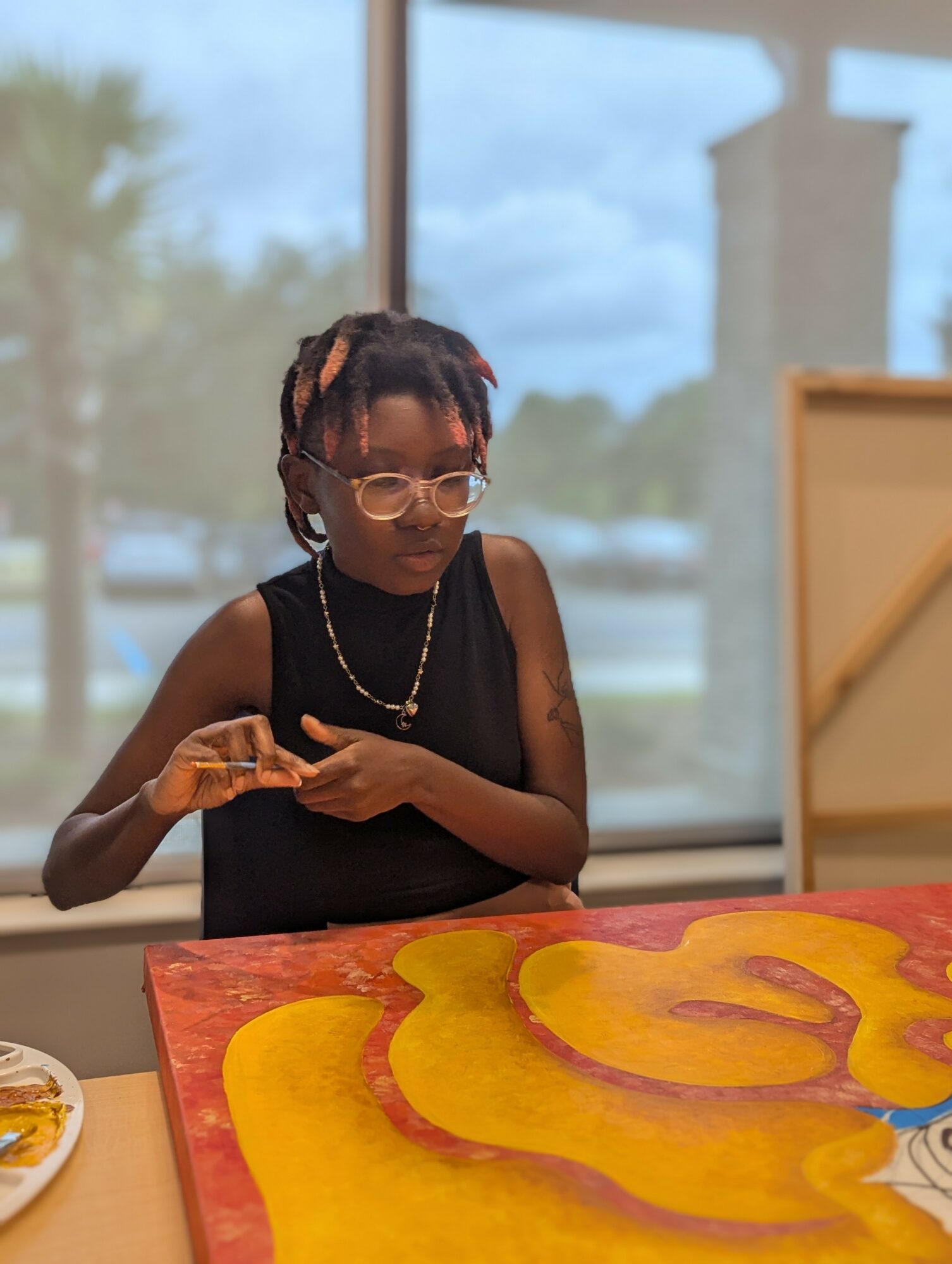 Young person with glasses and jewelry, sitting at a table with colorful artwork, in front of a window showing outdoor scenery.