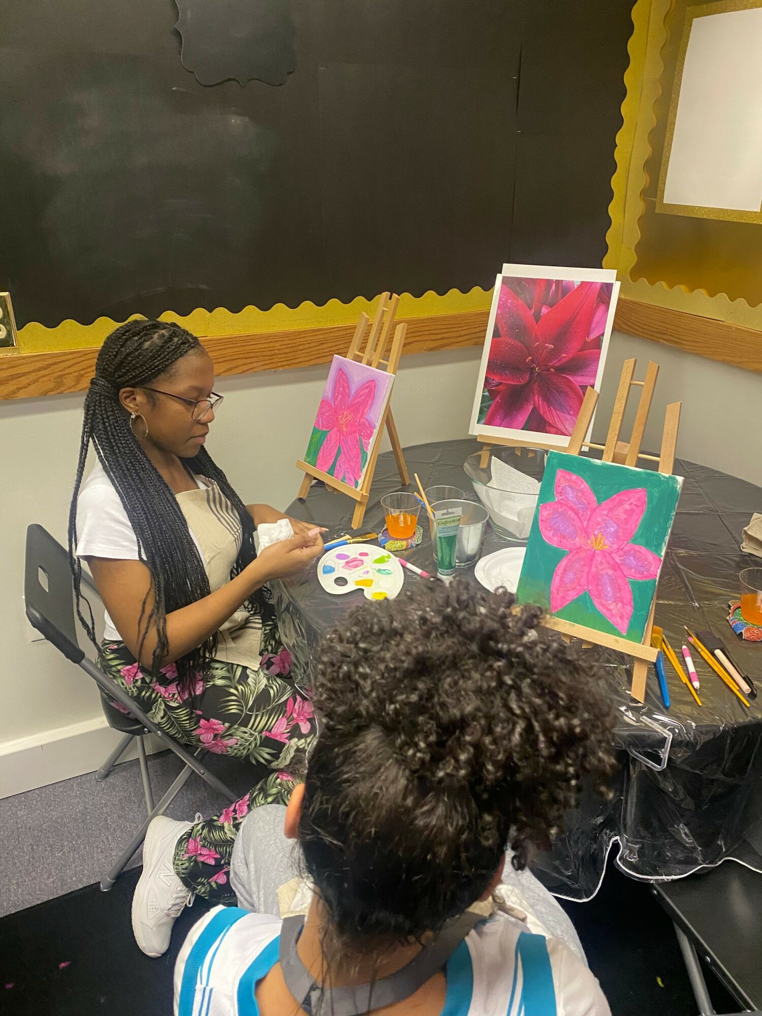 Child with curly hair painting a pink flower on paper, surrounded by colorful artwork and art supplies.