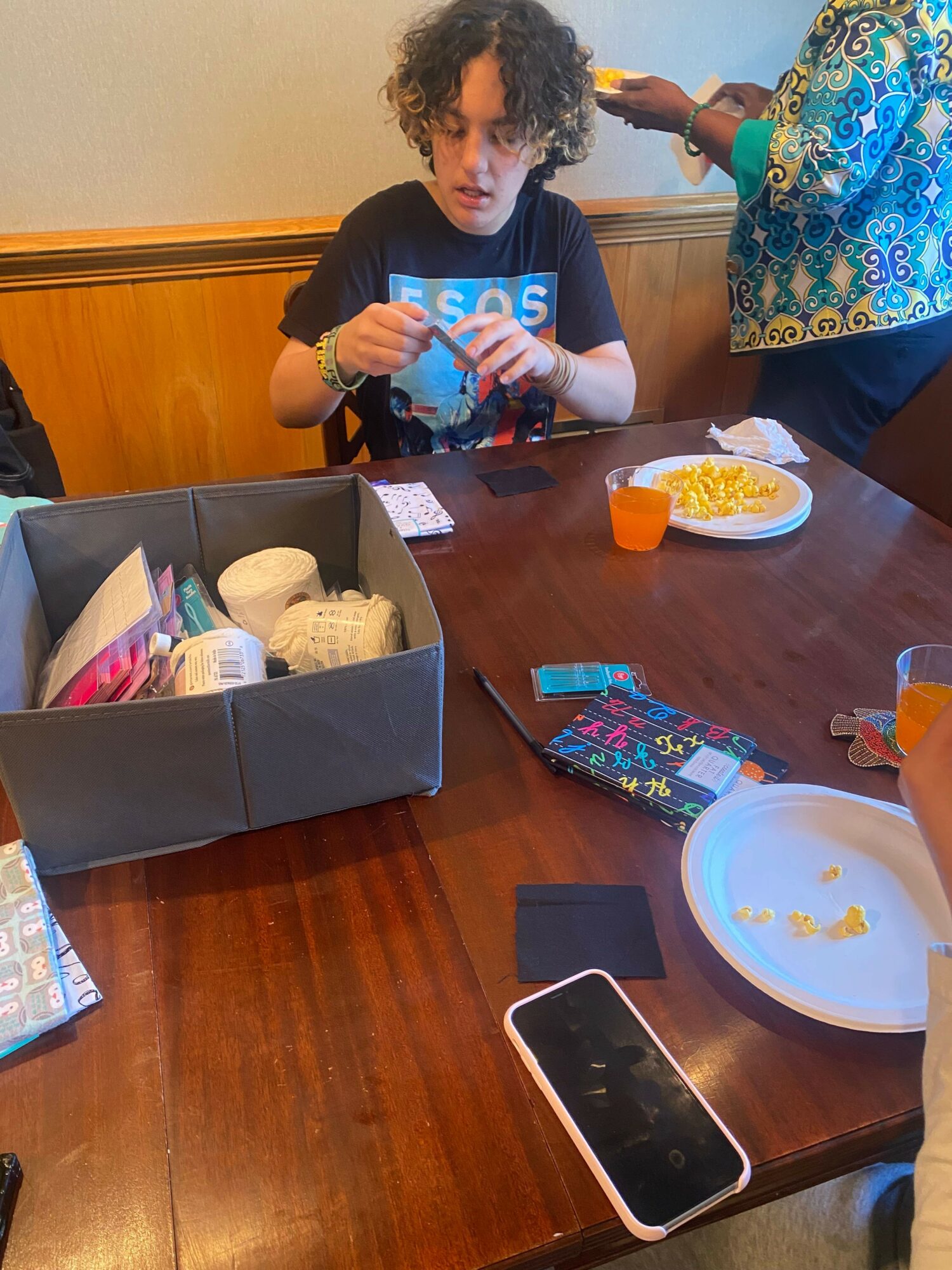 Child with curly hair and a blue shirt sitting at a table with plates, snacks, and a smartphone.