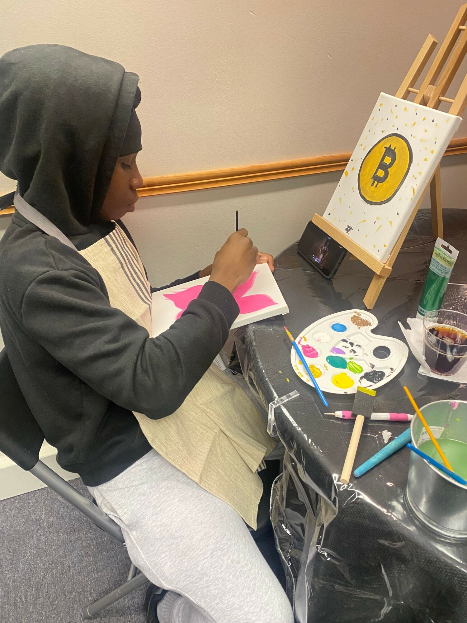 Person sitting at a table with art supplies, painting on paper, with a yellow and black logo on a stand nearby.