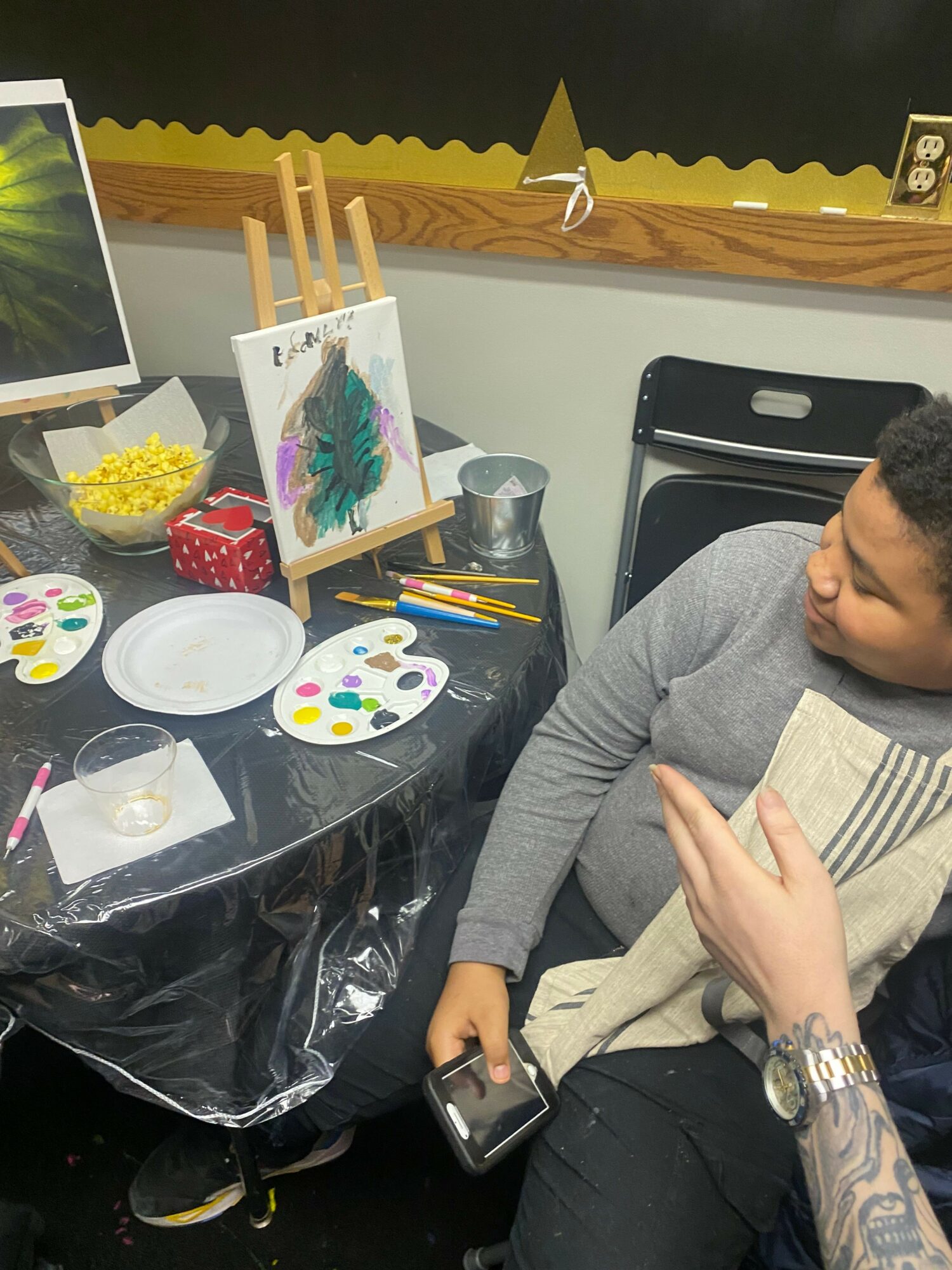 Person sitting at table with painting supplies, including paint palette, brushes, and a canvas with colorful artwork.