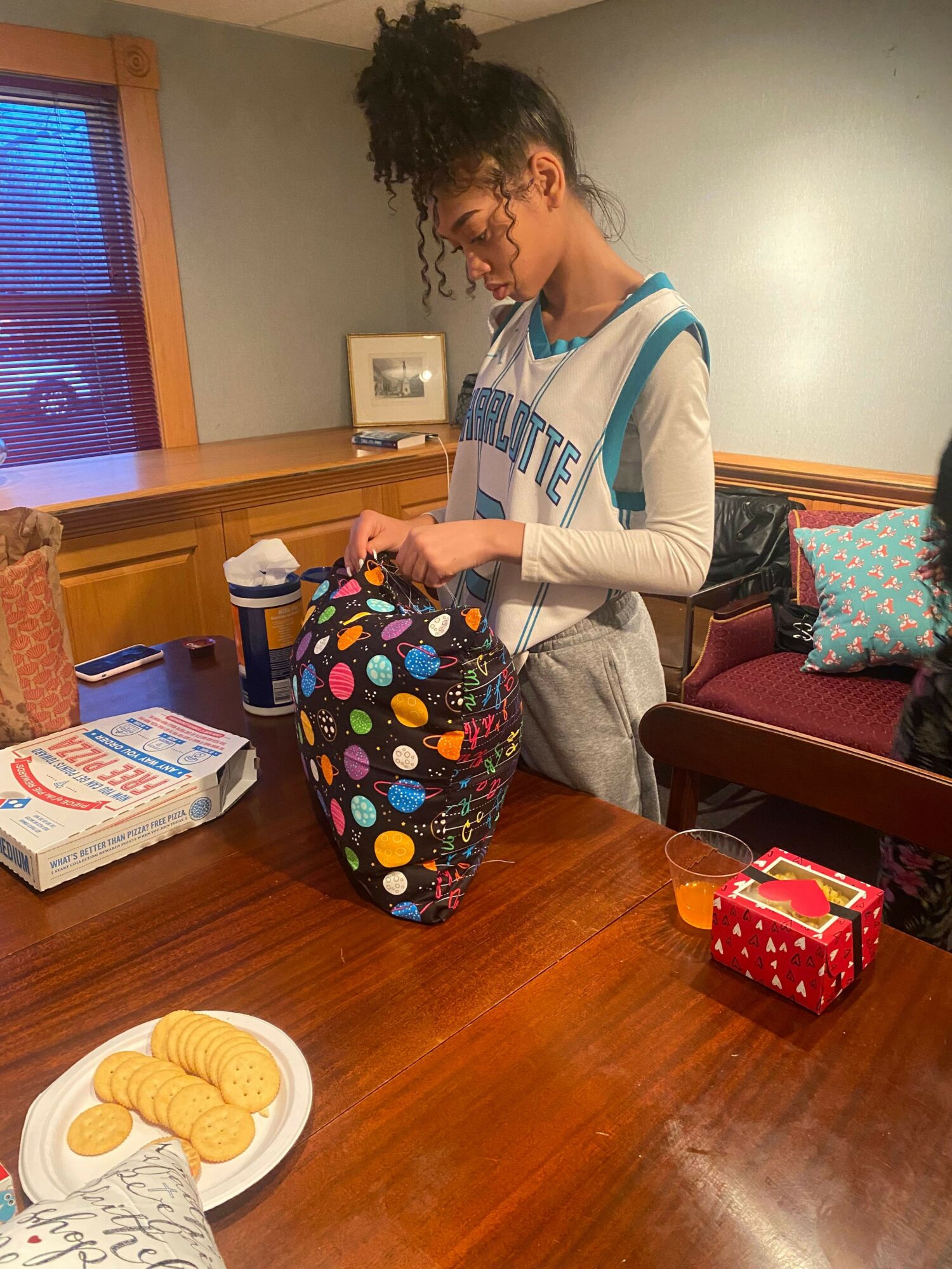 Child with curly hair in a white and blue shirt reaches into a colorful bag on a wooden table.