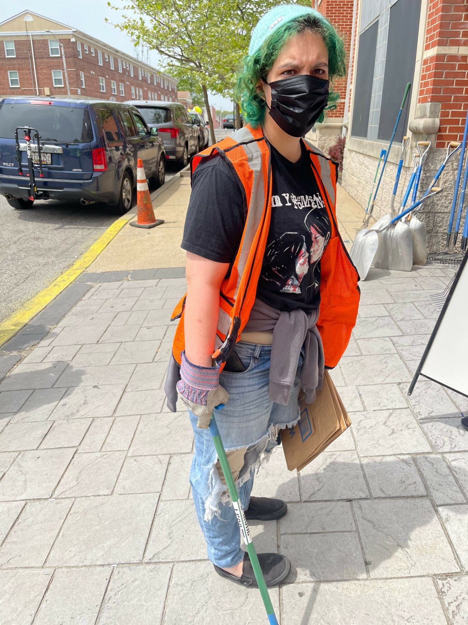 Person wearing a black mask, orange vest, and gray pants holding a broom on a sidewalk.