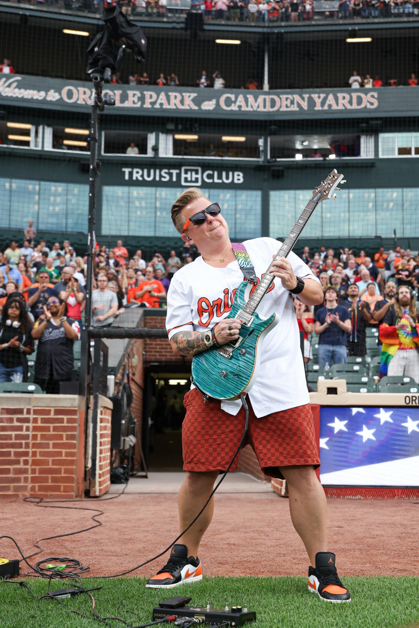 Person playing electric guitar on stage at Camden Yards with crowd in background, wearing sunglasses and casual clothing.