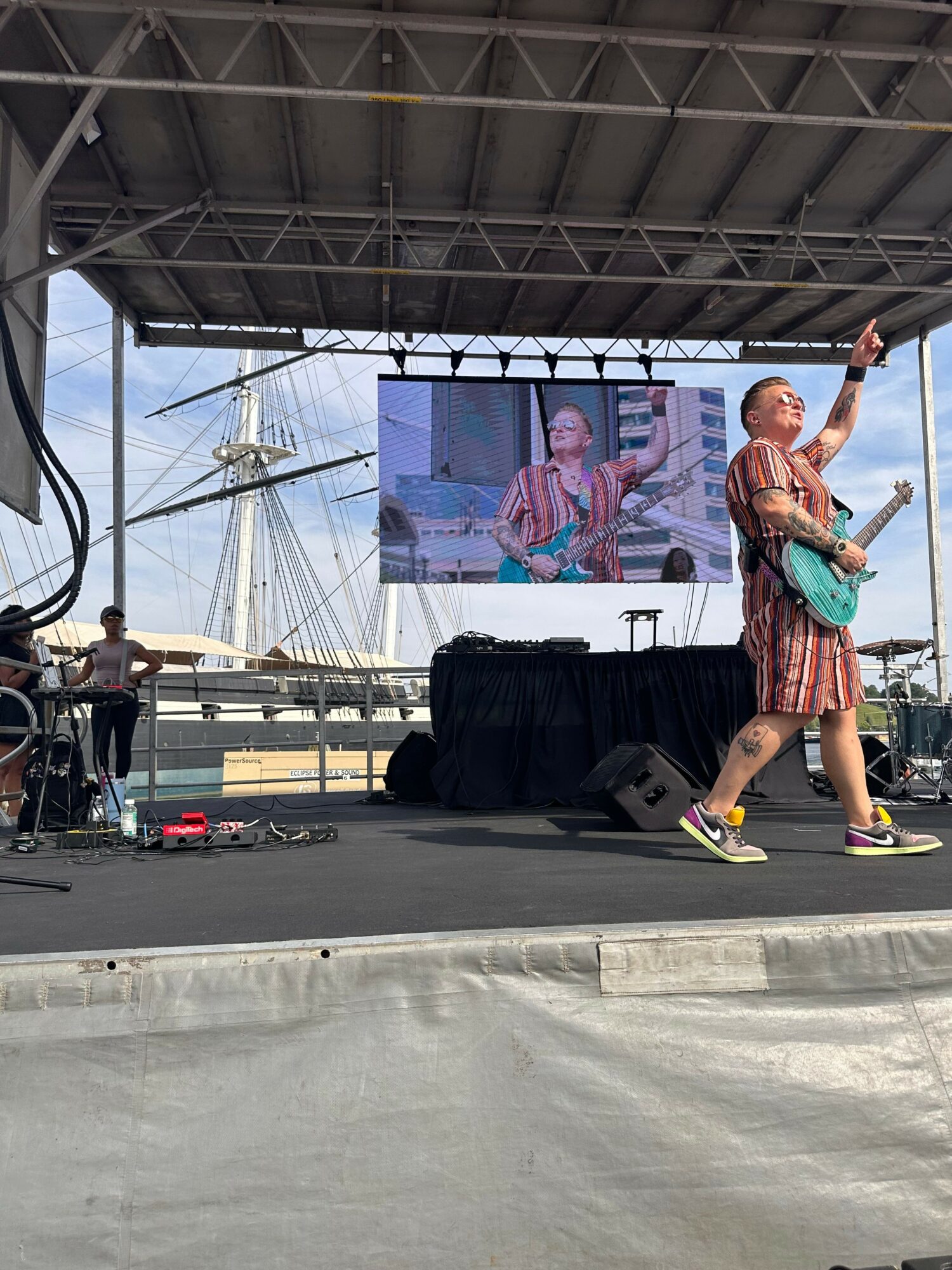 Person on stage holding a microphone, with a large screen and equipment behind, under a metal roof structure.
